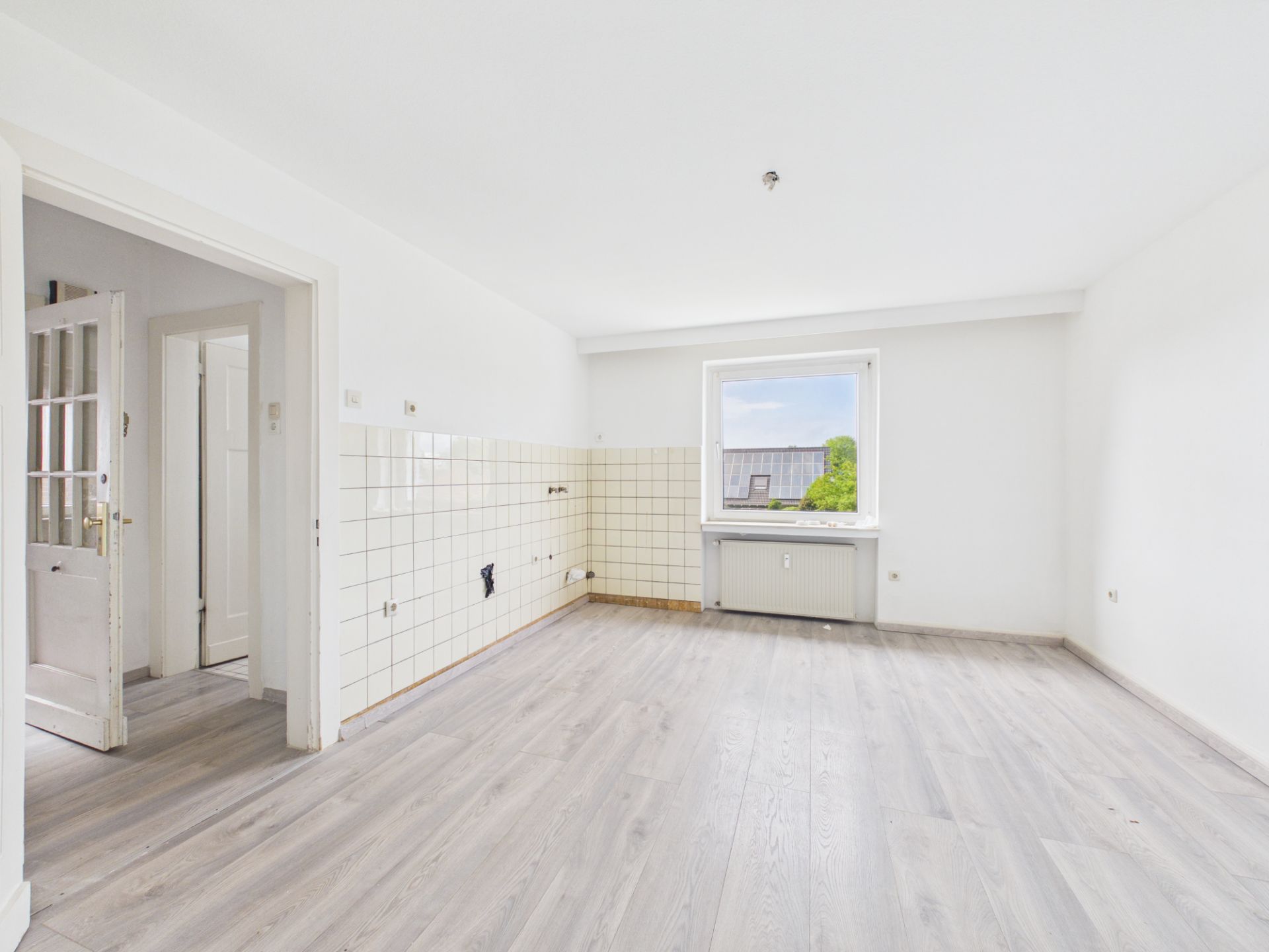 Leerer, heller Raum mit Fliesenwand, Laminatboden und Fenster mit Blick nach draußen.