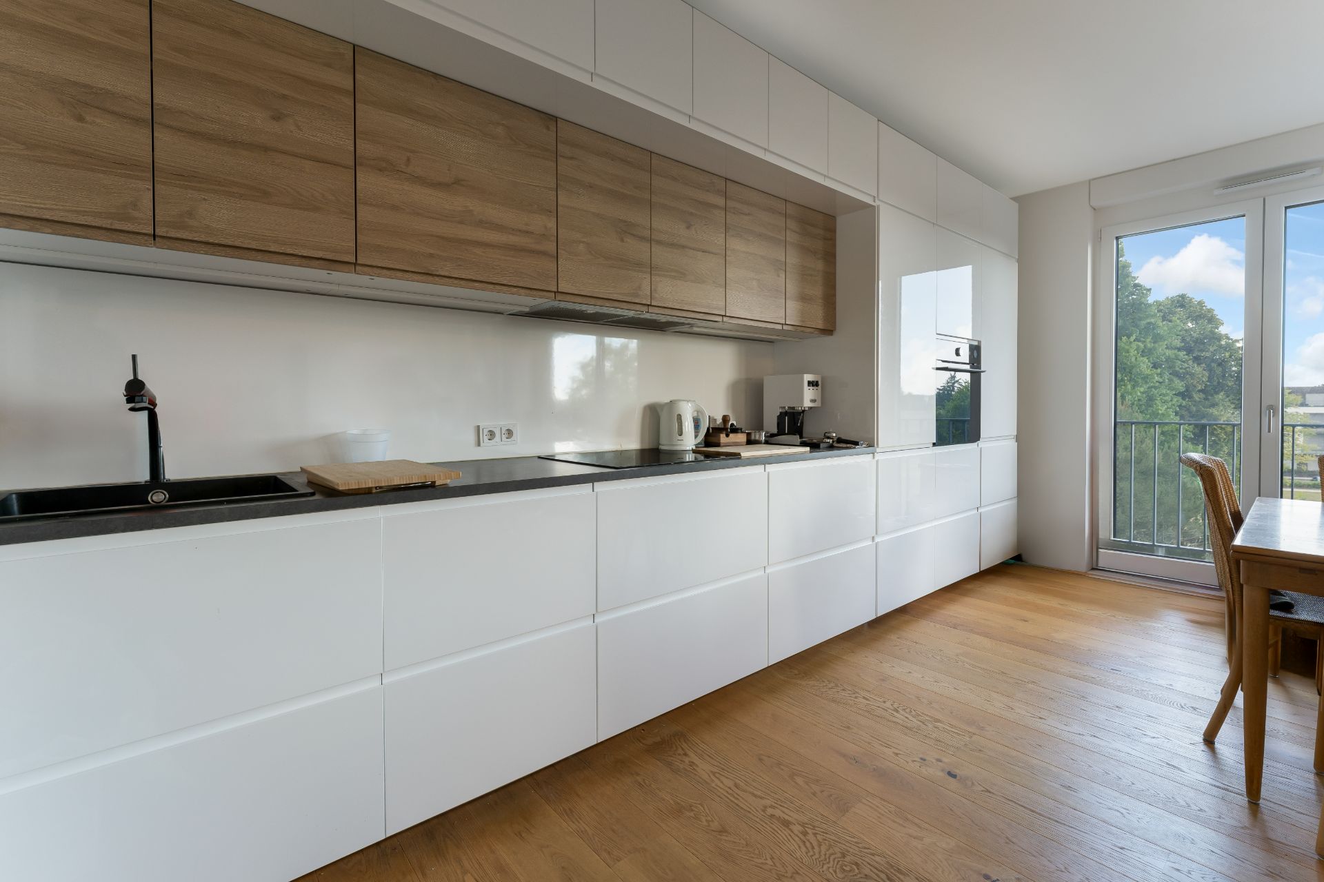 Moderne Küche mit weißen Schränken, Holzoberschränken und großem Fenster, Holzboden sichtbar.