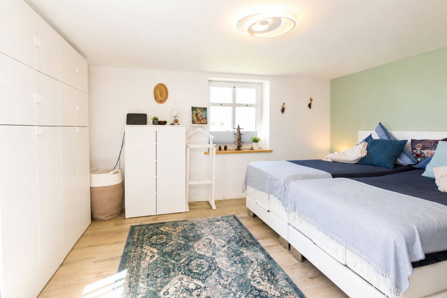 Helles Schlafzimmer mit zwei Betten, weißen Schränken und einem Fenster über einer Holzablage.