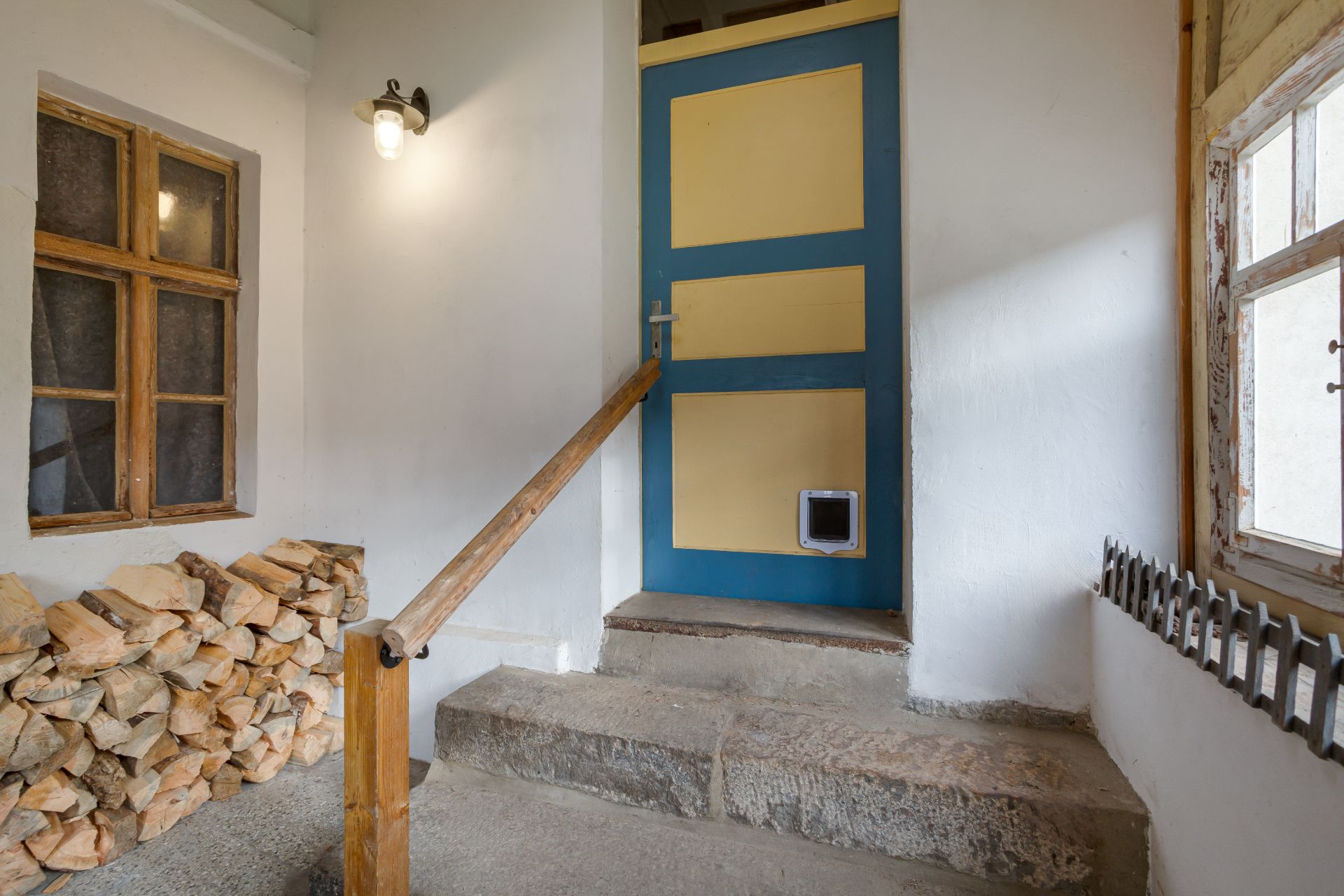 Eingangsbereich mit blauer Tür, Holzscheiten, zwei Fenstern und Treppe mit Holzgeländer.