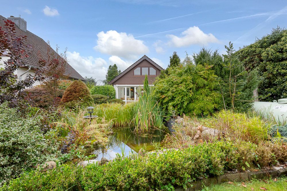 Garten mit großem Teich, vielen Pflanzen und Blick auf ein Wohnhaus im Hintergrund.