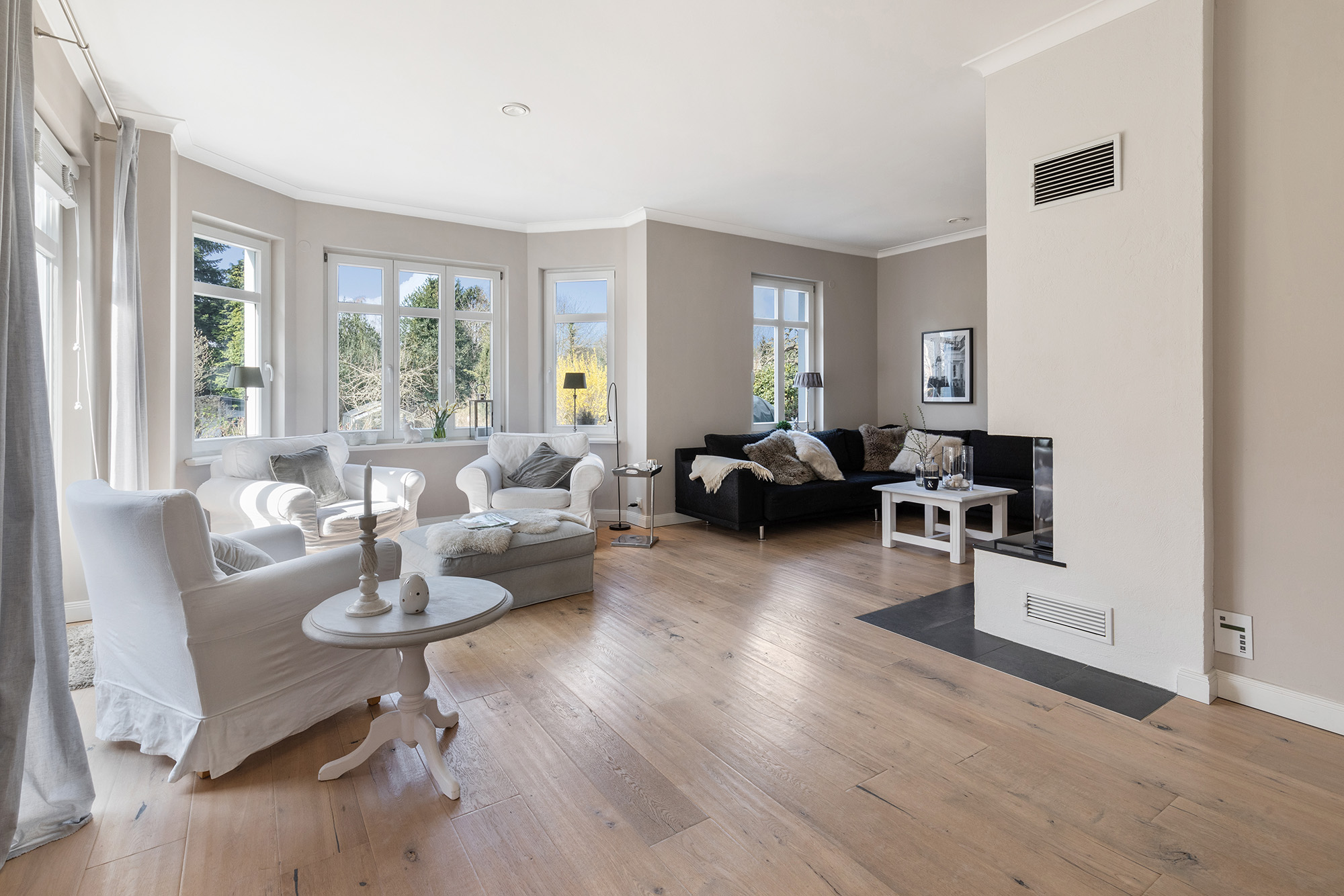 Helles Wohnzimmer mit großen Fenstern, hellen Sesseln und Sofa auf Holzfußboden.
