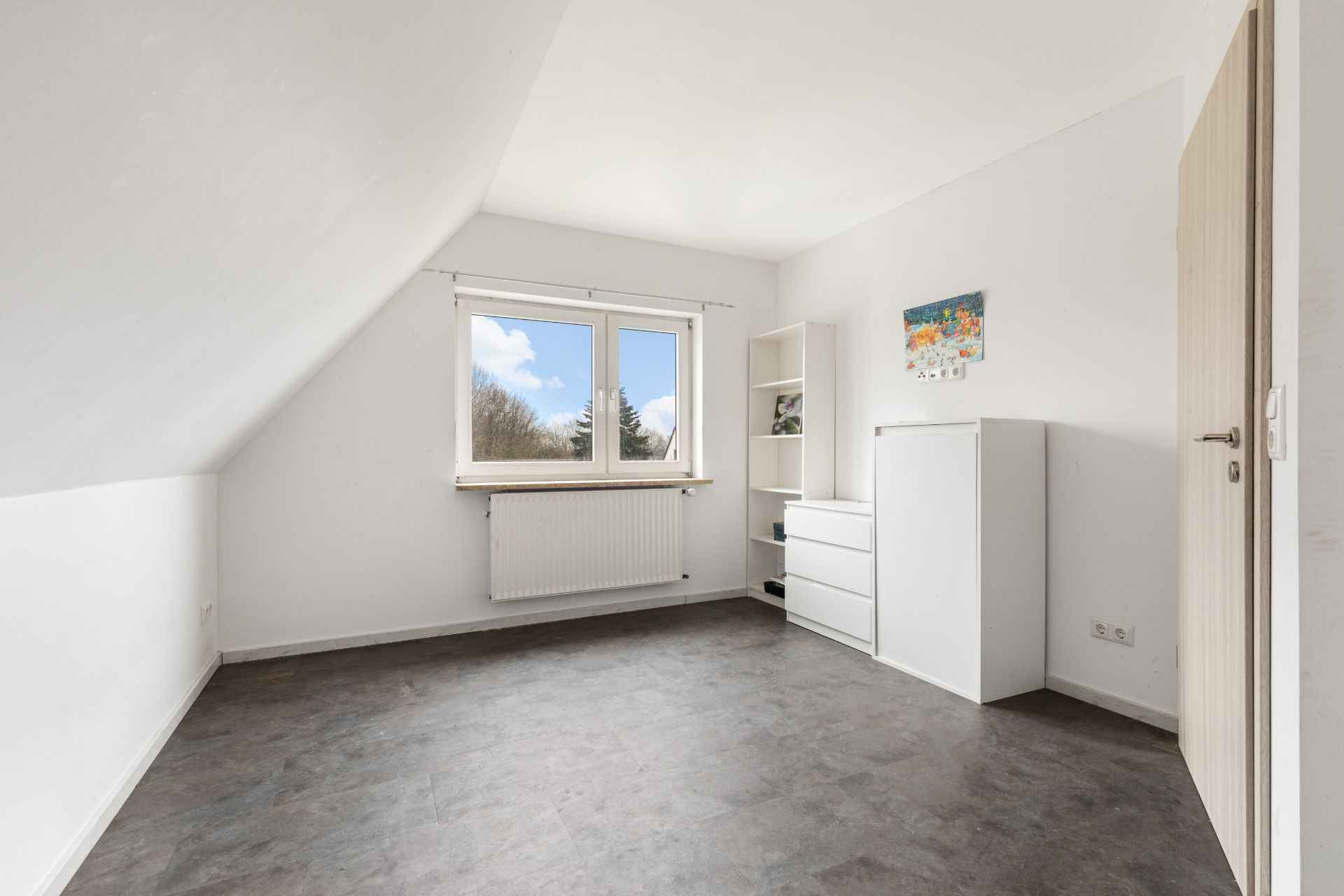 Helles Zimmer mit Dachschräge, Fenster, weißem Regal und Kommode auf dunklem Boden.