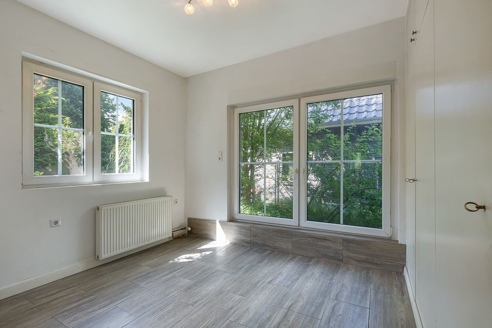 Heller Raum mit großen Fenstern, Heizung und Fliesenboden, Blick ins Grüne.