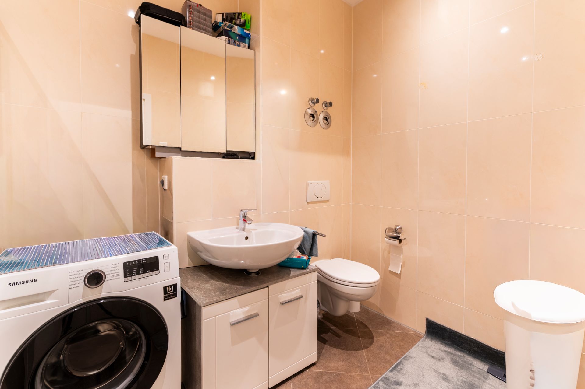 Modernes Badezimmer mit Waschmaschine, Waschbecken, Spiegel, Toilette und Mülleimer.