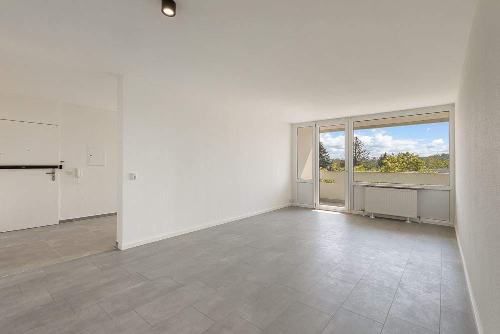 Leeres, helles Zimmer mit grauem Fliesenboden und großen Fenstern mit Ausblick ins Grüne.