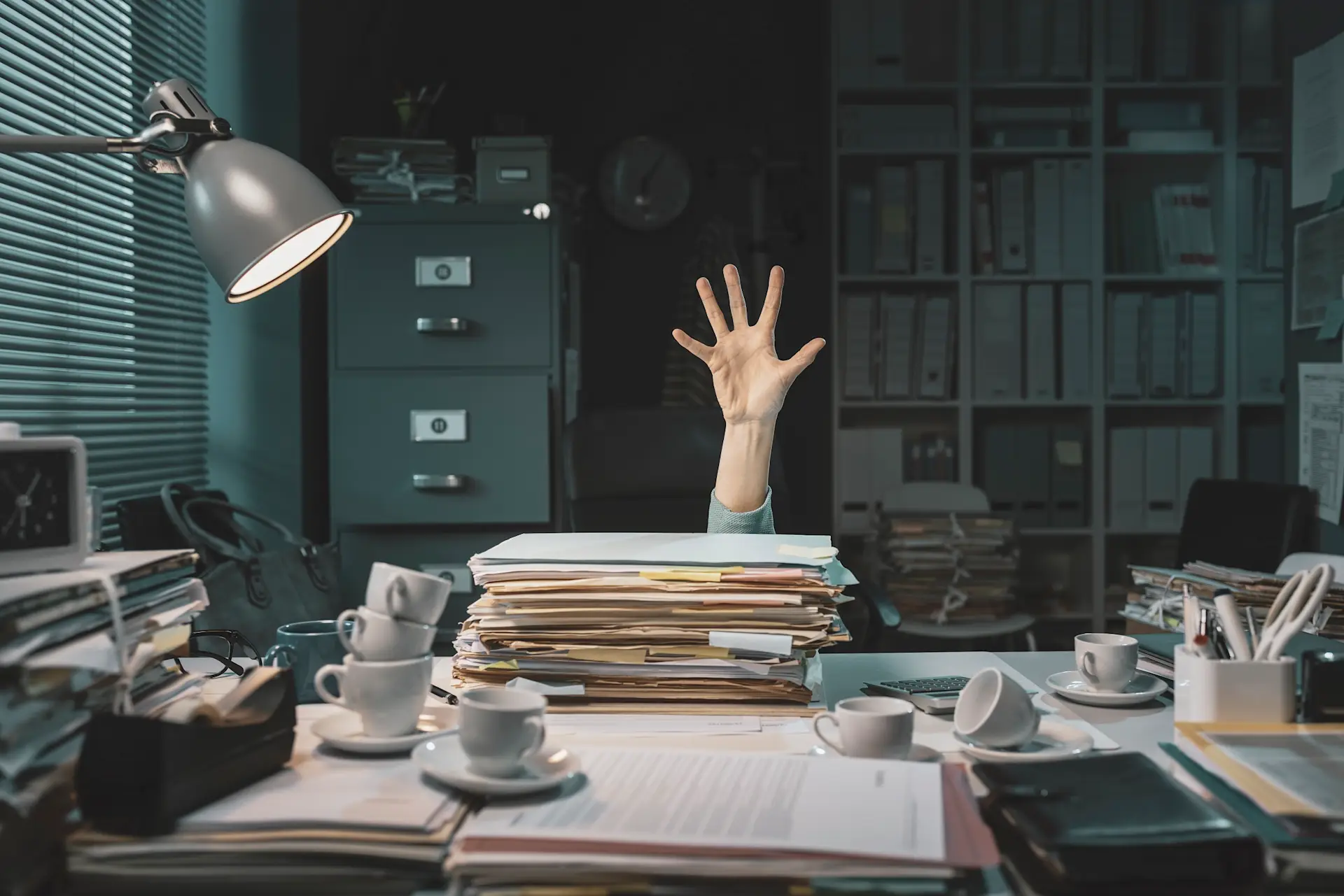 Hand ragt aus einem Stapel Akten auf einem überfüllten Schreibtisch im Büro.