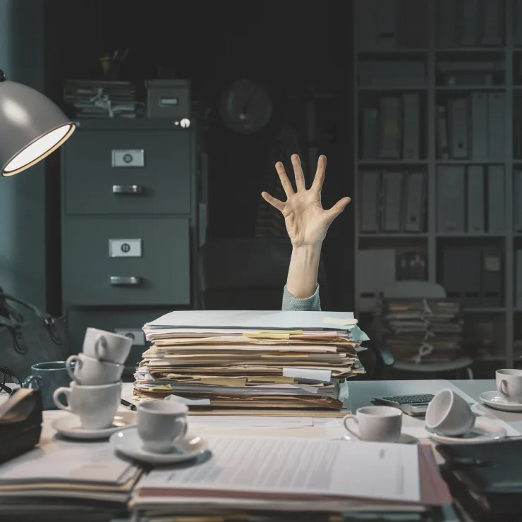 Hand ragt aus einem Stapel Akten auf einem überfüllten Schreibtisch im Büro.
