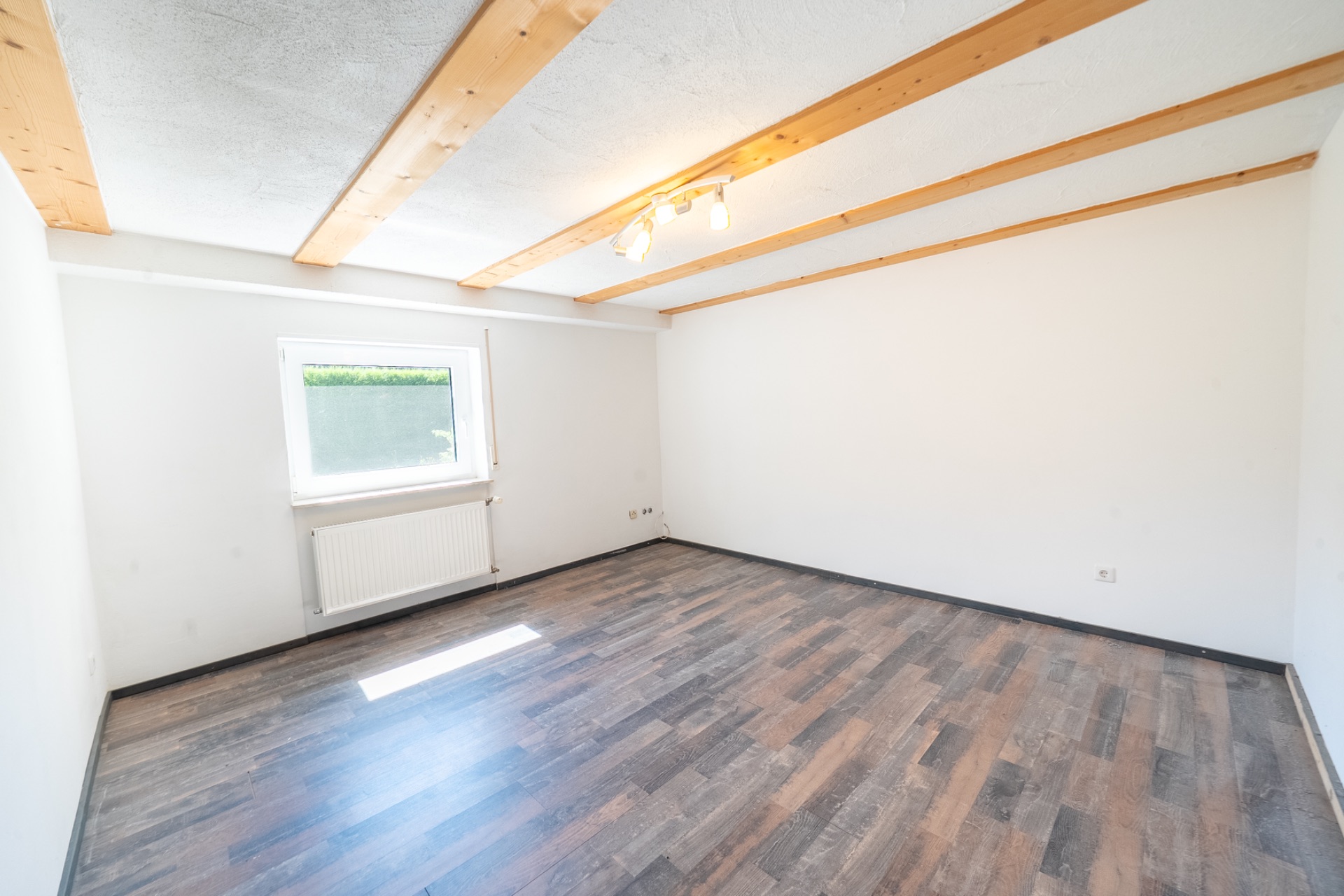 Leeres Zimmer mit Holzbalkendecke, Fenster, Heizung und dunklem Laminatboden.
