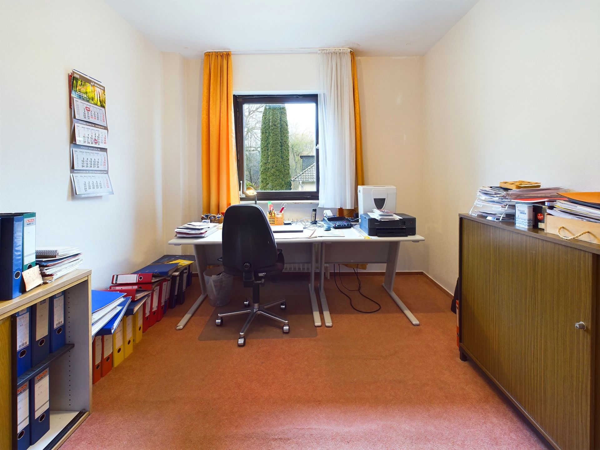 Helles Arbeitszimmer mit Schreibtisch, Stuhl, Regalen und Fenster mit gelbem Vorhang.