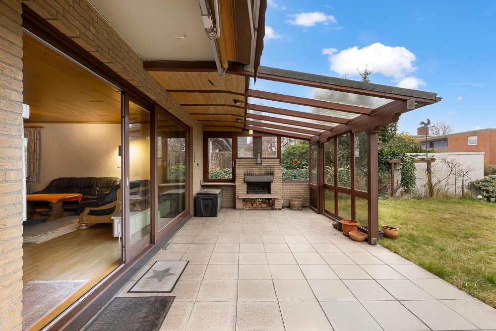 Überdachte Terrasse mit Glaswänden, Grill und Blick in den Garten.