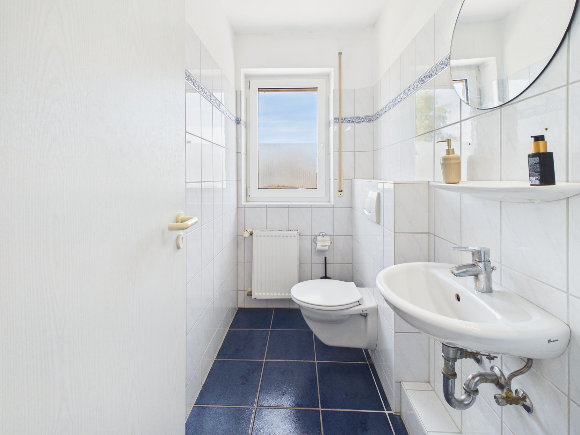 Helles Badezimmer mit blauem Fliesenboden, Wand-WC, Waschbecken und Fenster.