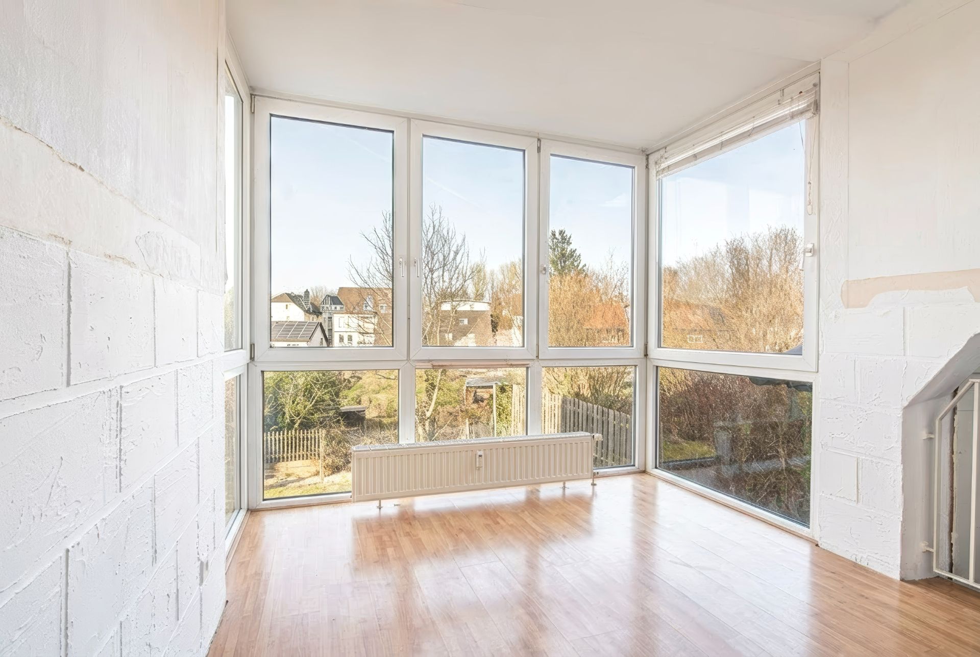 Heller Raum mit großen Fenstern, Parkettboden und Blick auf einen Garten.
