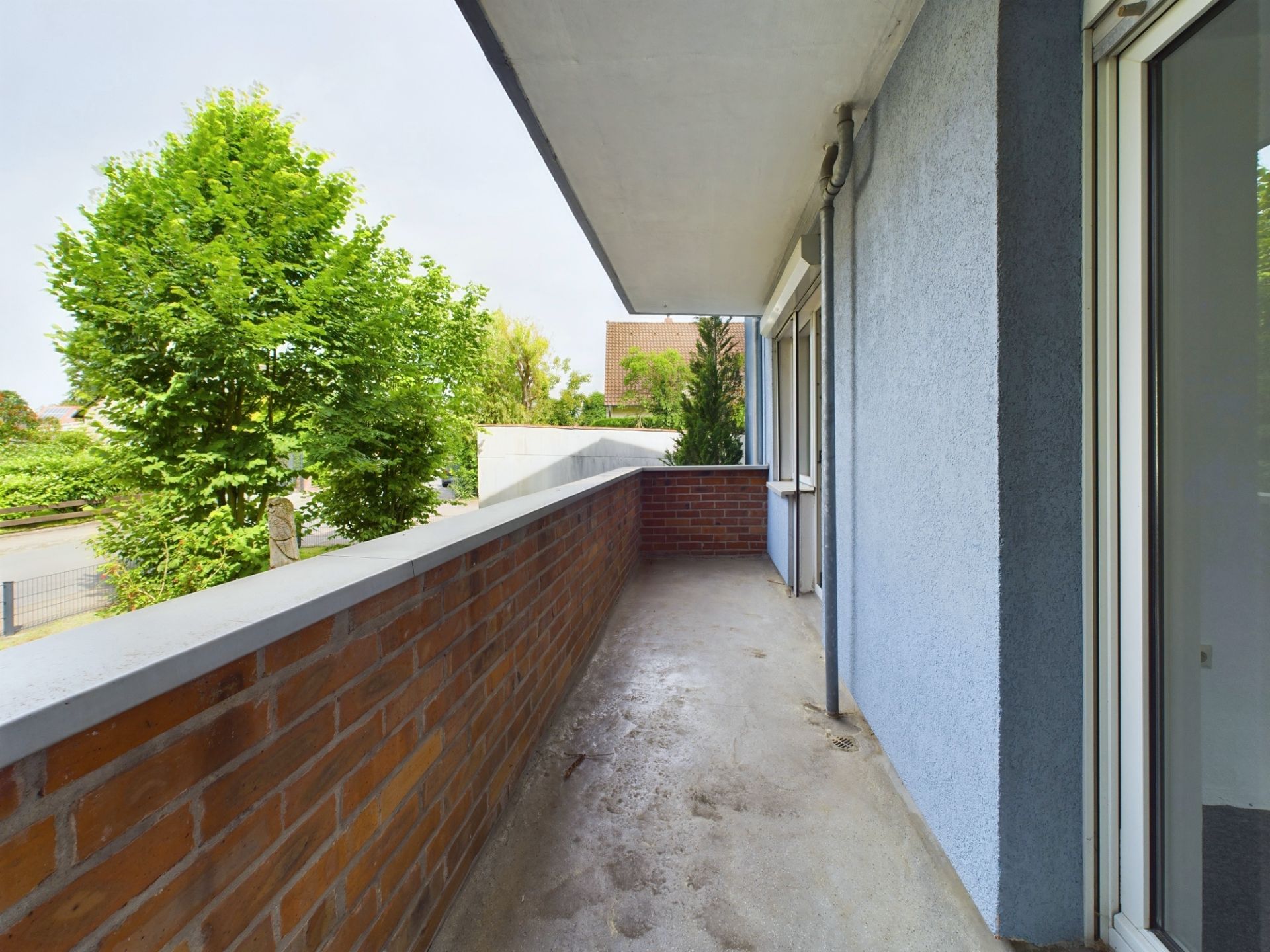 Langer Balkon mit Backsteinbrüstung, Blick auf grüne Bäume und angrenzende Straße.