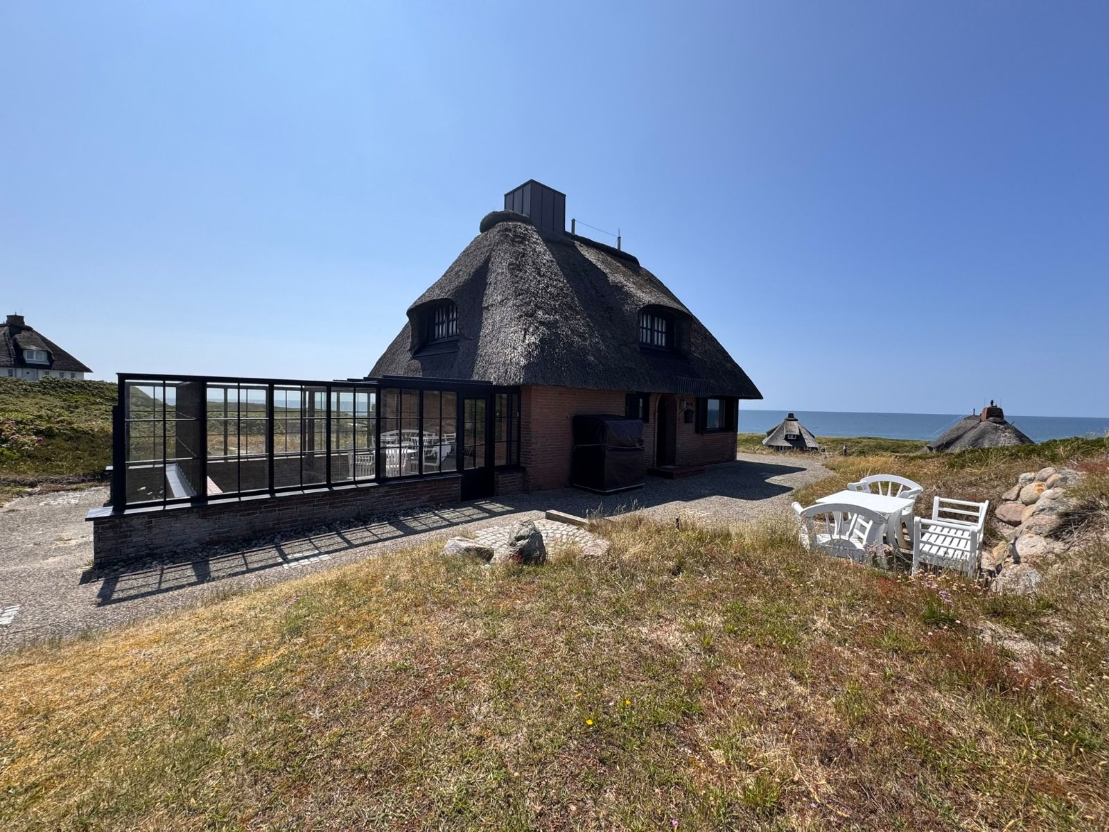 Reetgedecktes Haus mit Wintergarten, Gartenmöbeln und Blick aufs Meer bei klarem Himmel.