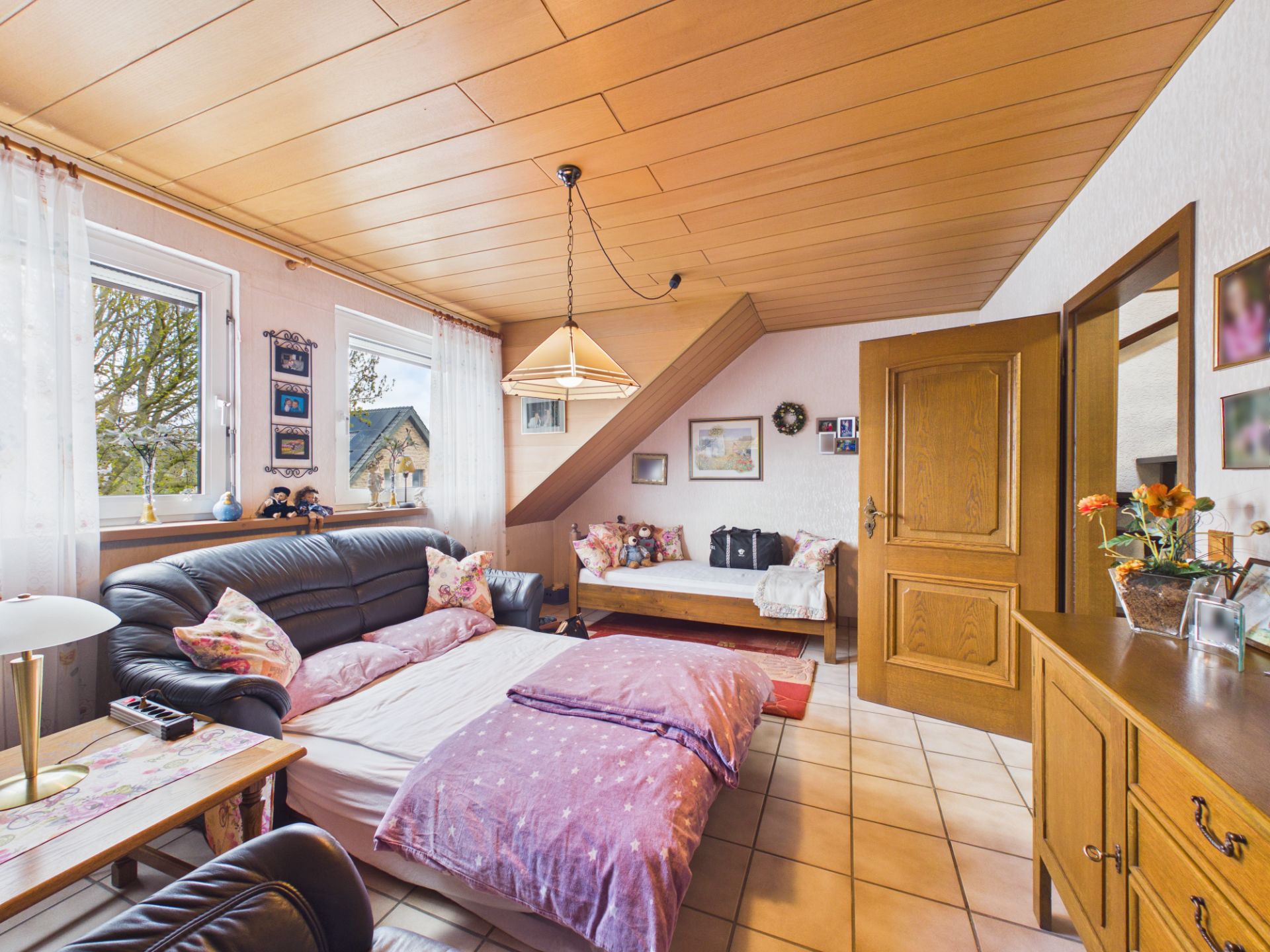 Wohnzimmer mit Schlafsofa, Einzelbett, Holzmöbeln und Fenster mit Vorhängen.