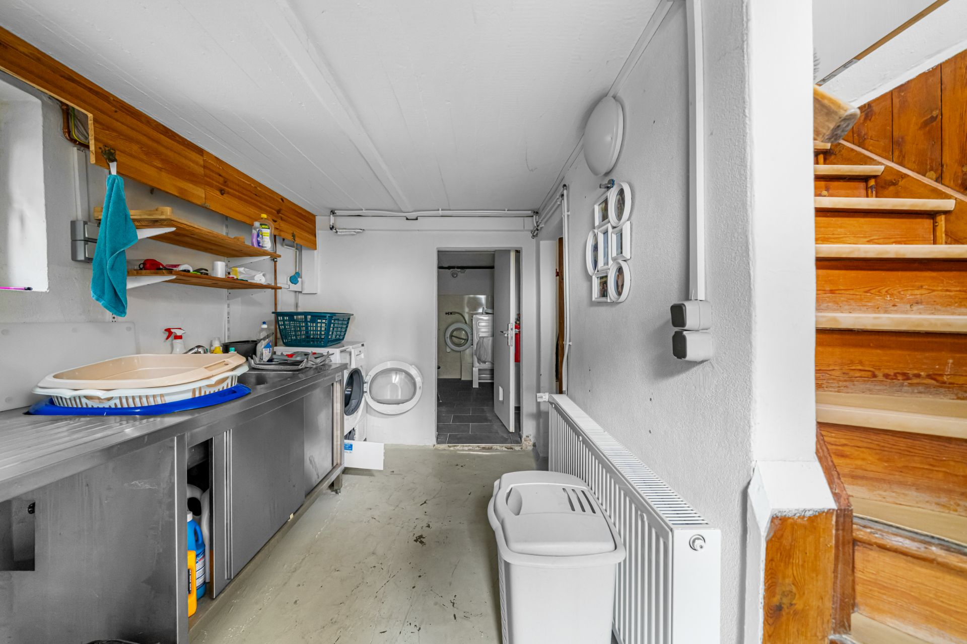 Waschküche mit Waschmaschine, Regal, Waschbecken und Holztreppe am rechten Bildrand.
