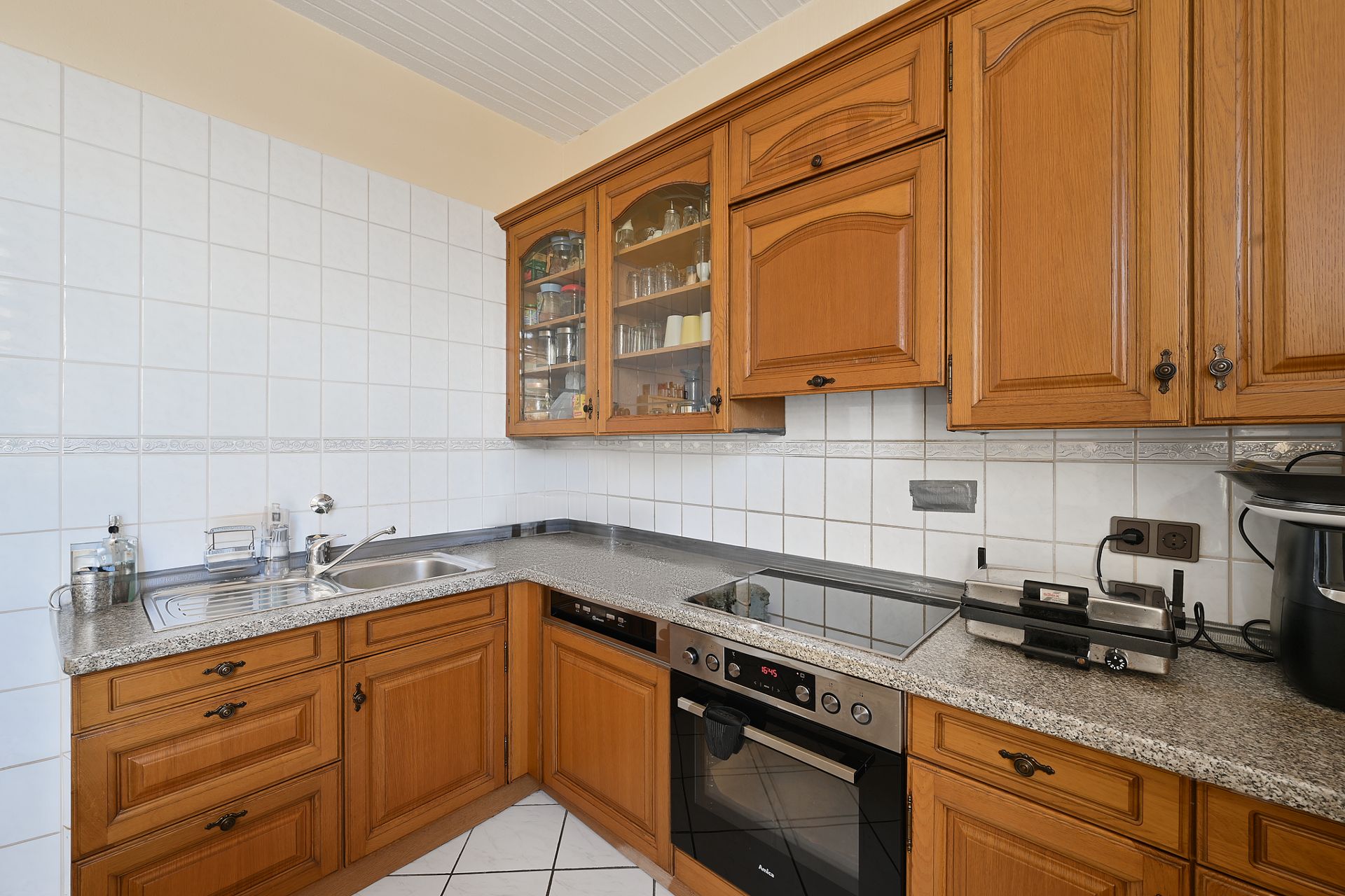 Eckküche mit Holzschränken, Ceranfeld, Backofen und Spüle, weiße Fliesen an der Wand.