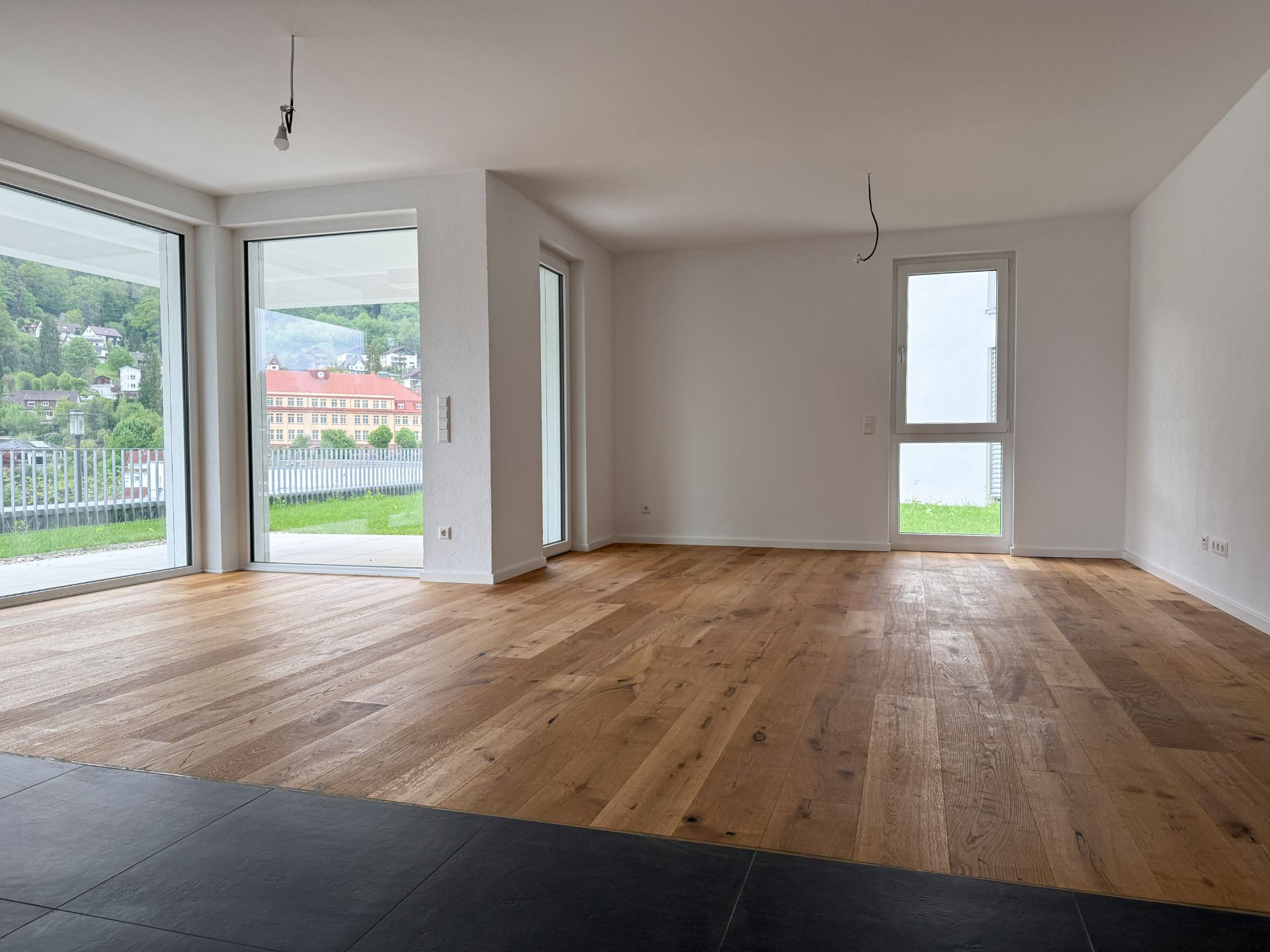Helles, leeres Wohnzimmer mit großem Holzfußboden und bodentiefen Fenstern.