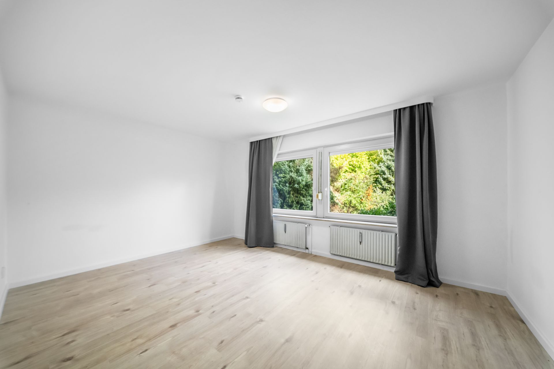 Helles, leeres Zimmer mit Holzfußboden, großem Fenster und grauen Vorhängen.