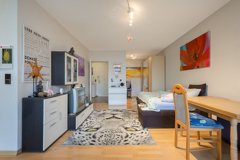 Helles Wohnzimmer mit Teppich, Bett, Esstisch, Kommode und Wandbildern.