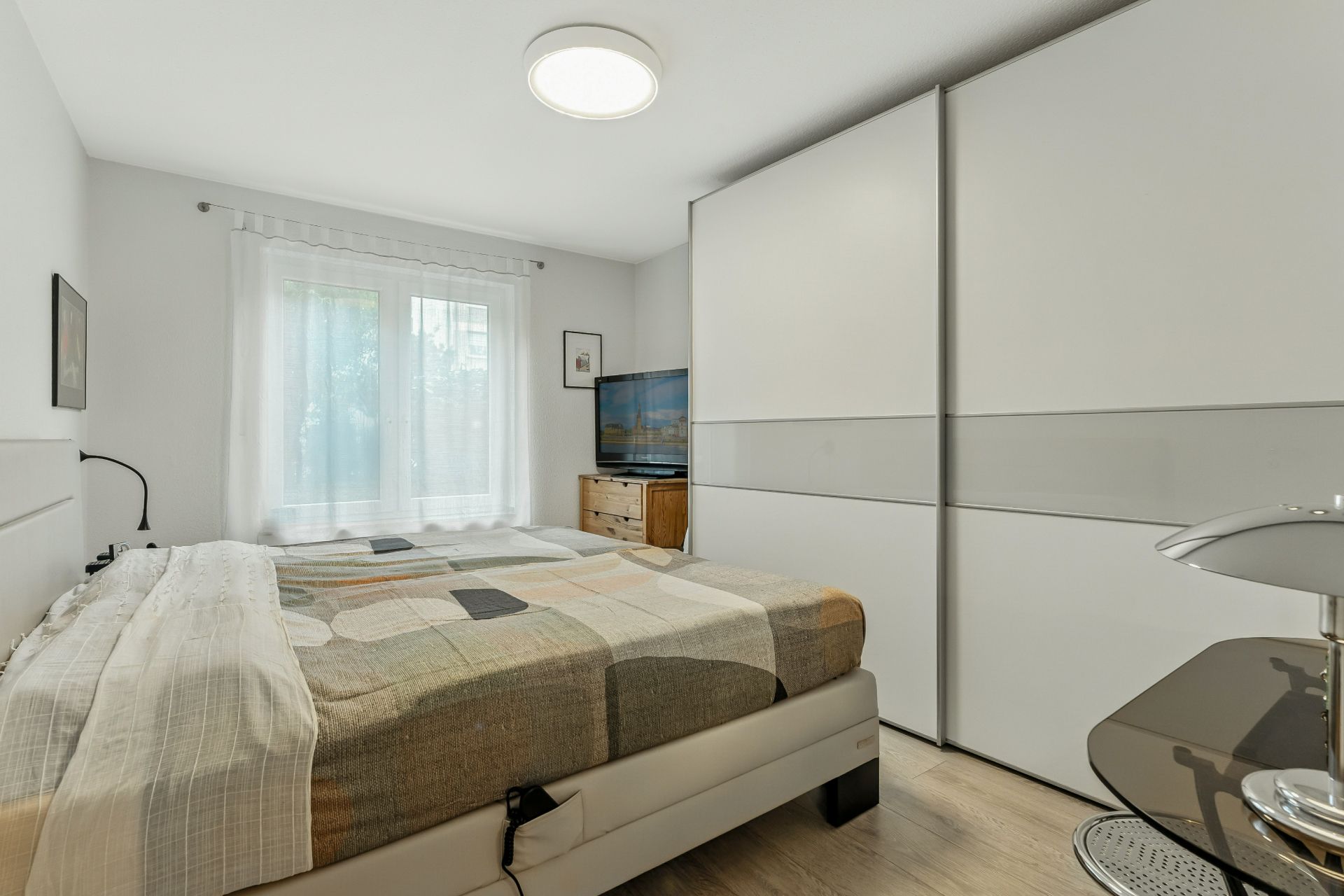 Helles Schlafzimmer mit Doppelbett, großem Schrank, Fenster und Fernseher auf Kommode.
