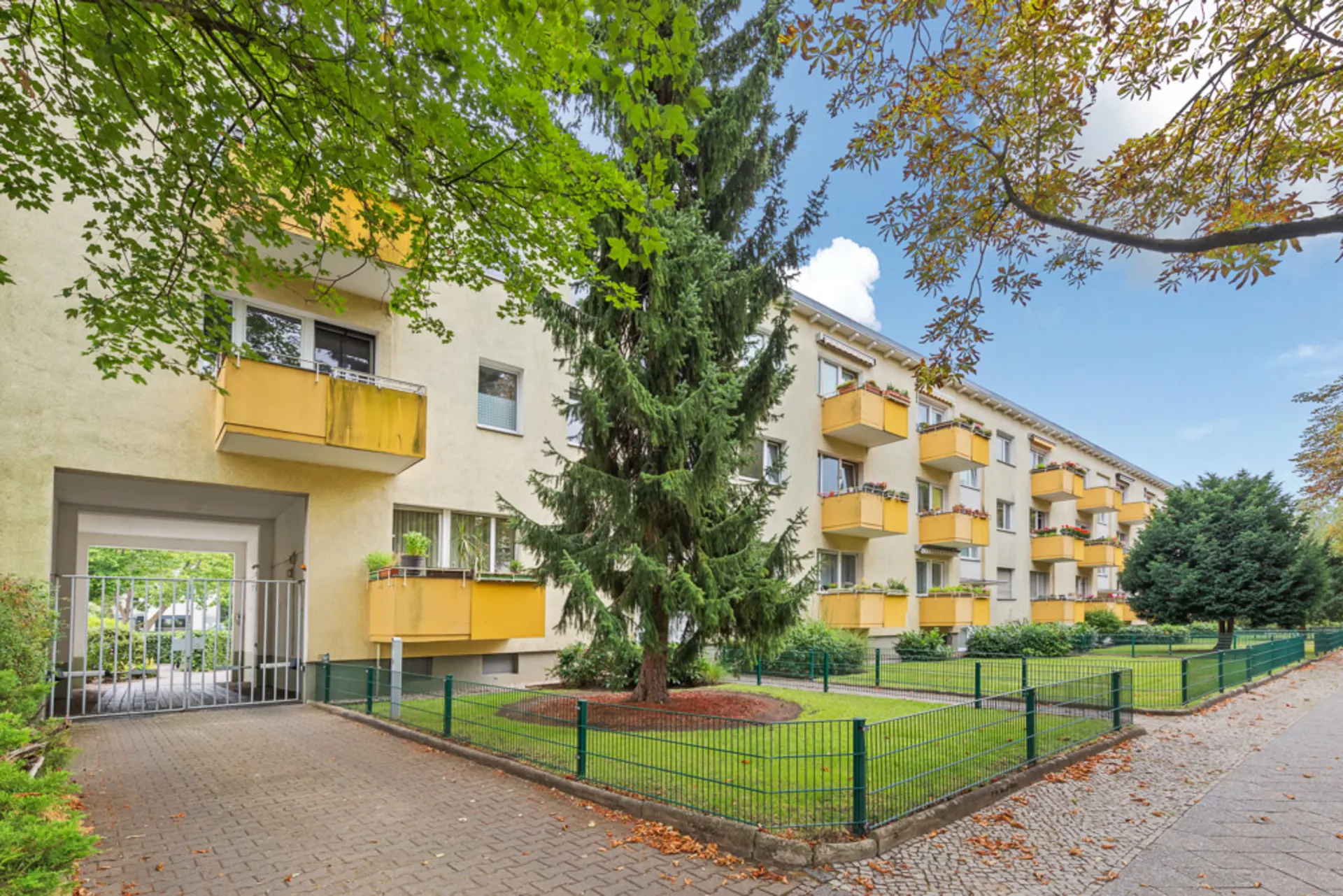 Mehrfamilienhaus mit gelben Balkonen, grünem Vorgarten und Einfahrtstor.