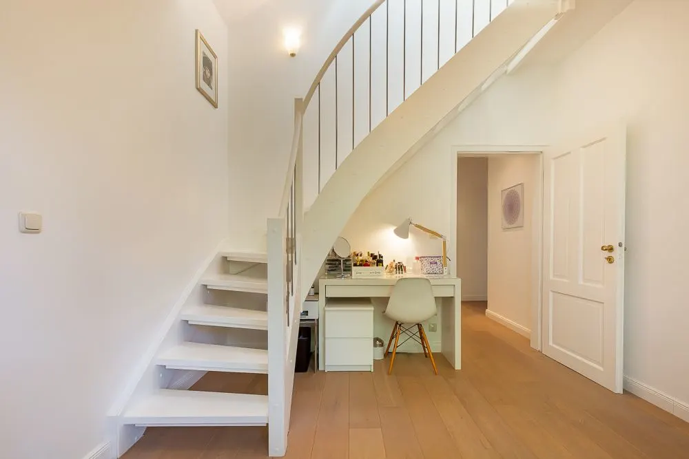 Weiße Treppe mit Arbeitsbereich darunter, Schreibtisch, Stuhl und Lampe in hellem Raum.