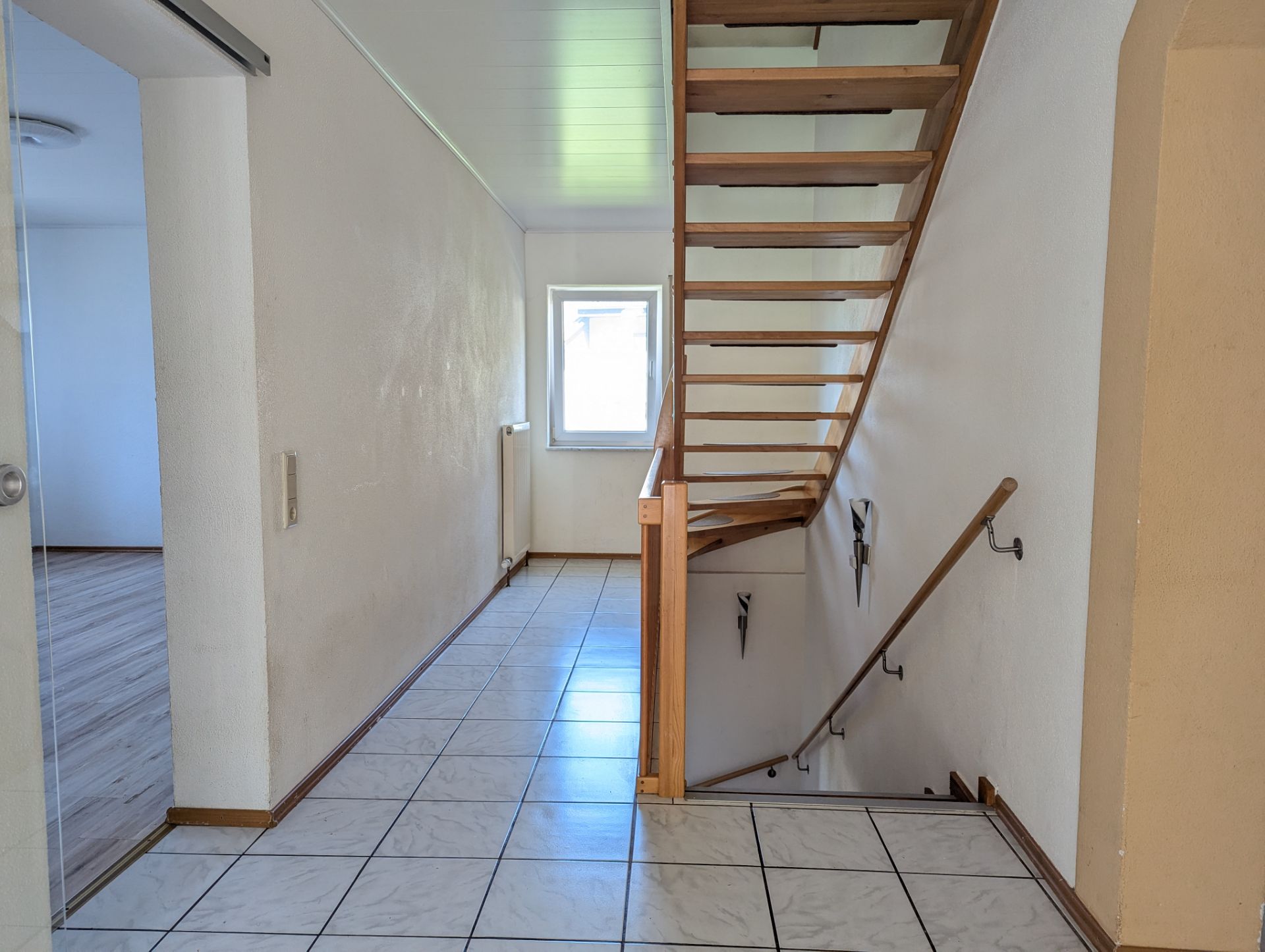 Flur mit gefliestem Boden und offener Holztreppe, Fenster am Ende des Gangs.