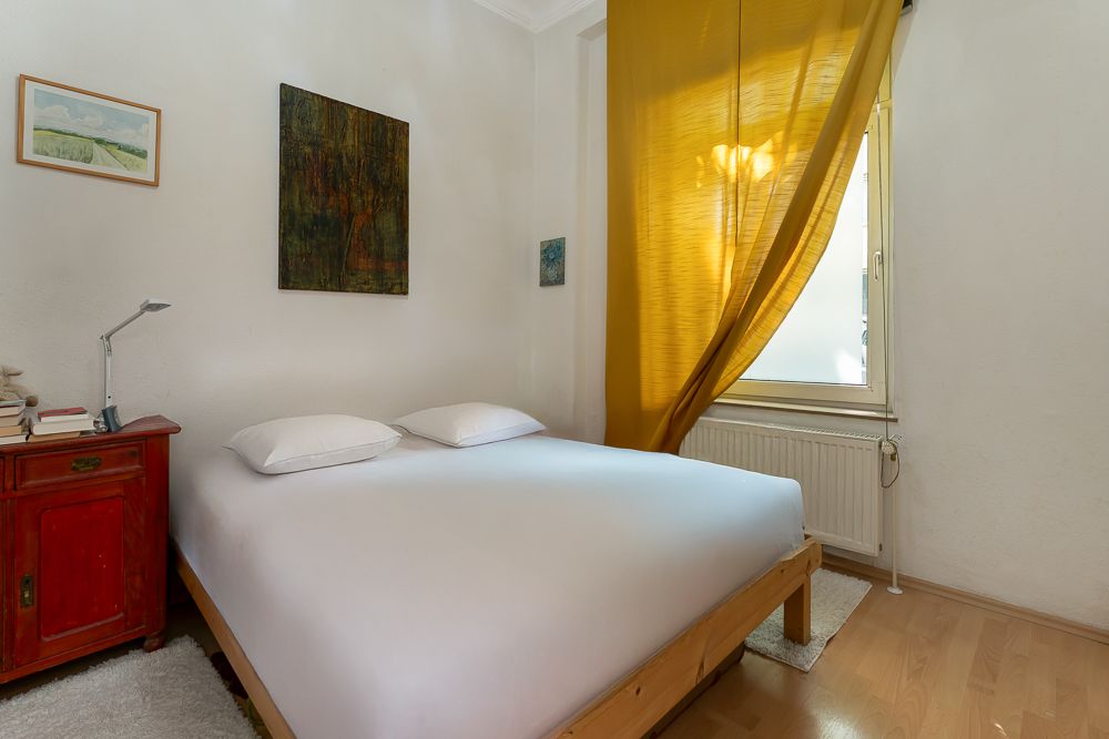 Schlafzimmer mit Doppelbett, rotem Nachttisch und gelbem Vorhang am Fenster.