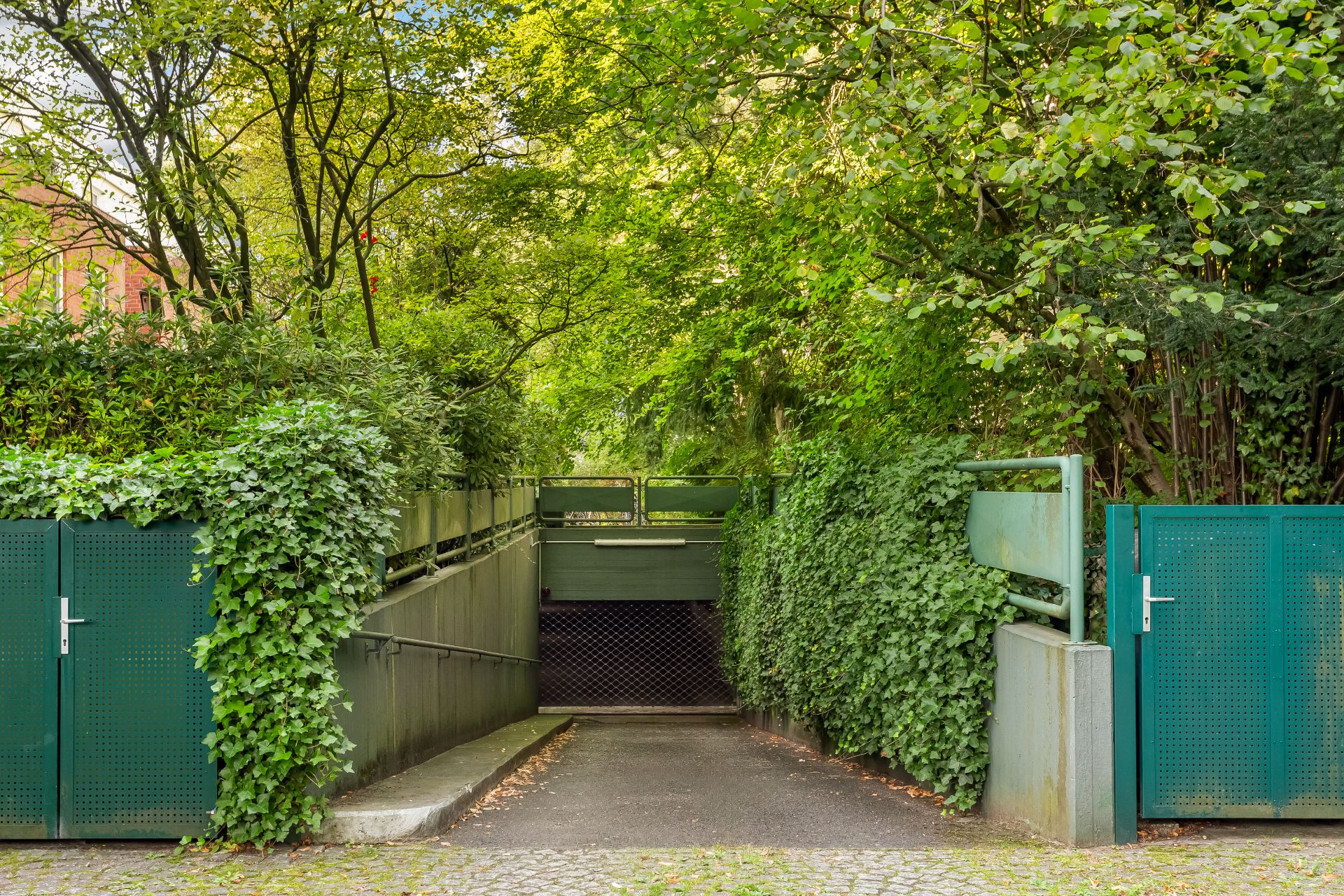 Einfahrt zu einer Tiefgarage mit grünen Toren und viel Bewuchs rundherum.