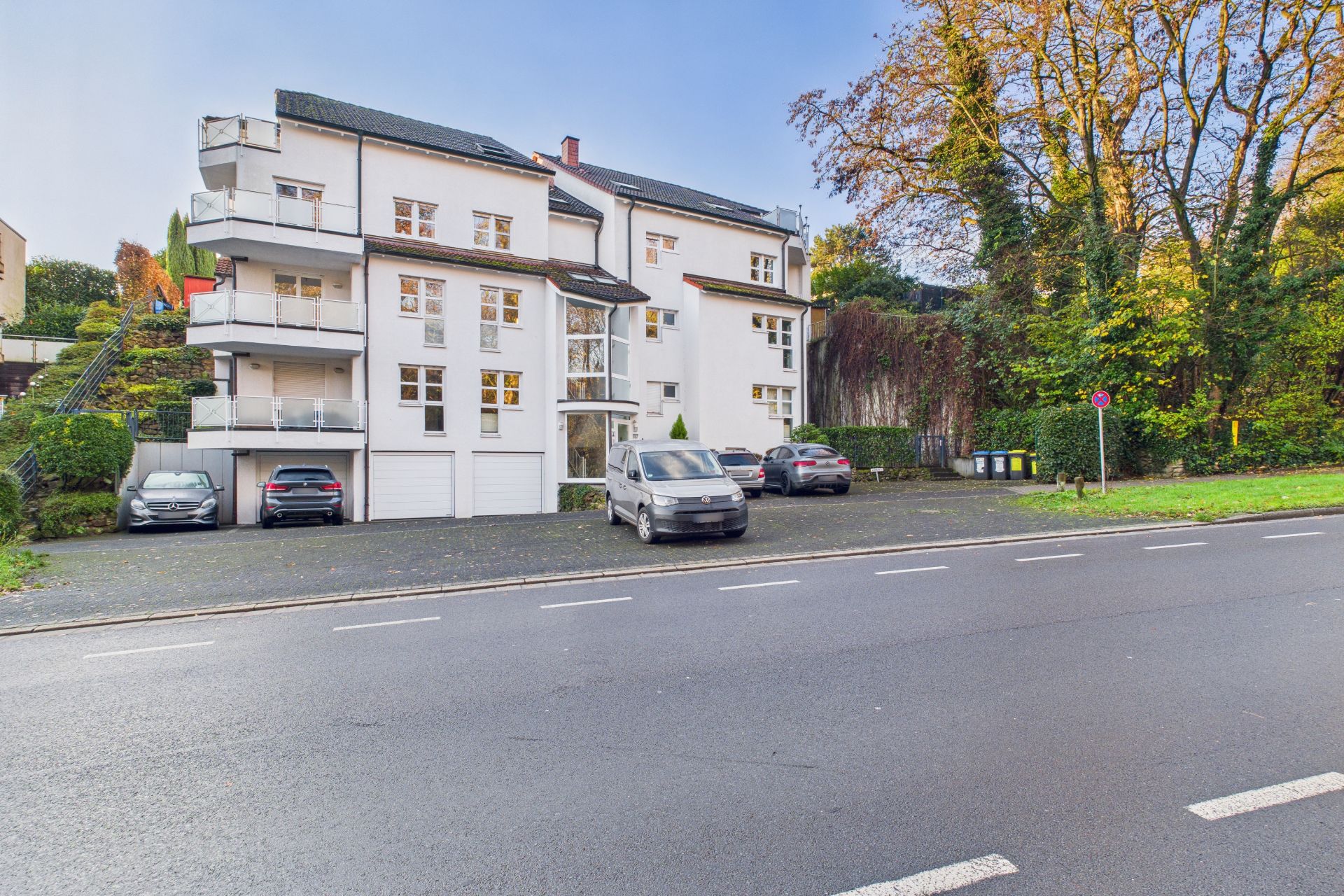 Weiße Wohnhausfassade mit mehreren Balkonen und parkenden Autos davor an einer Straße.