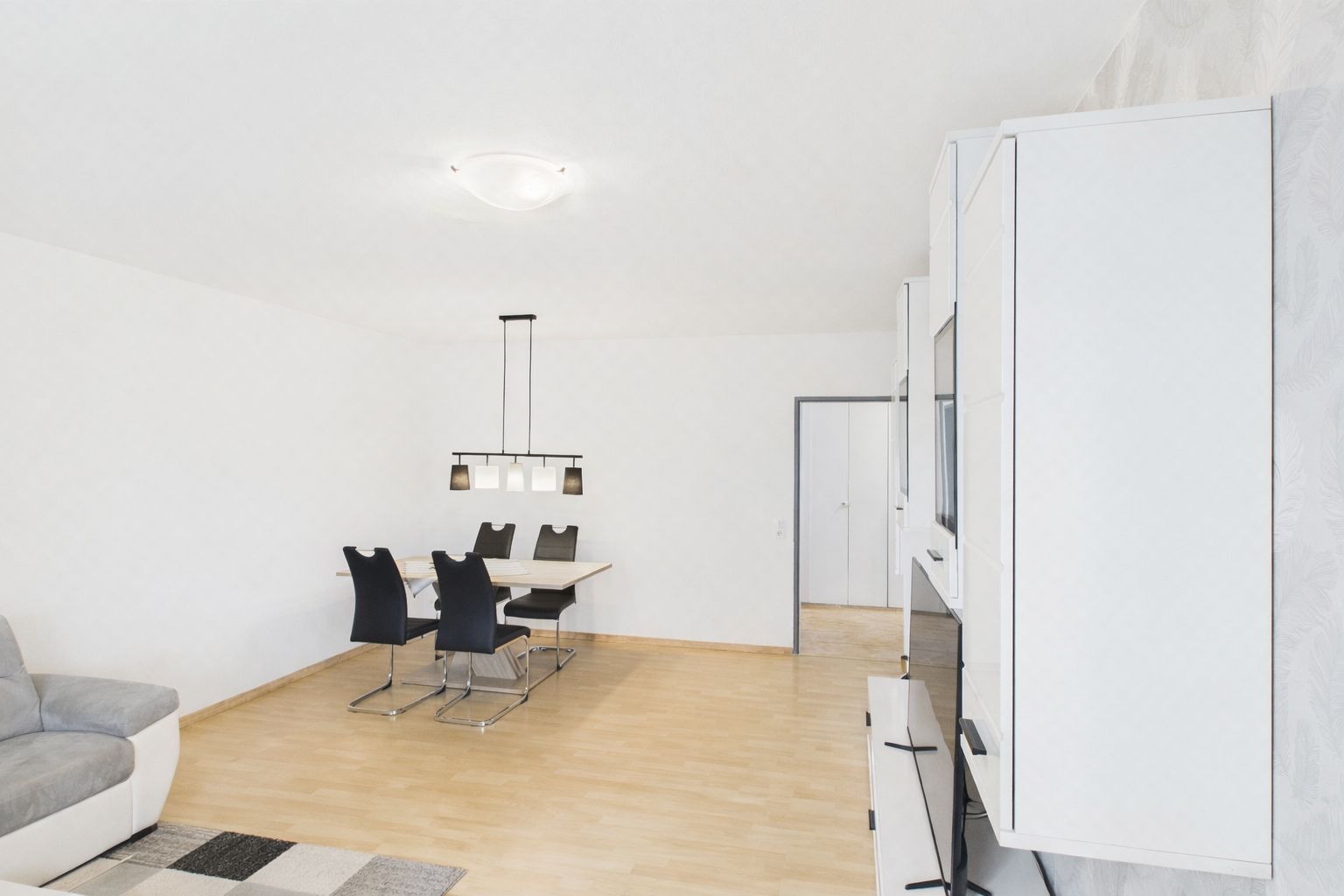 Helles Wohnzimmer mit Esstisch, vier schwarzen Stühlen und weißem Schrank auf Parkettboden.