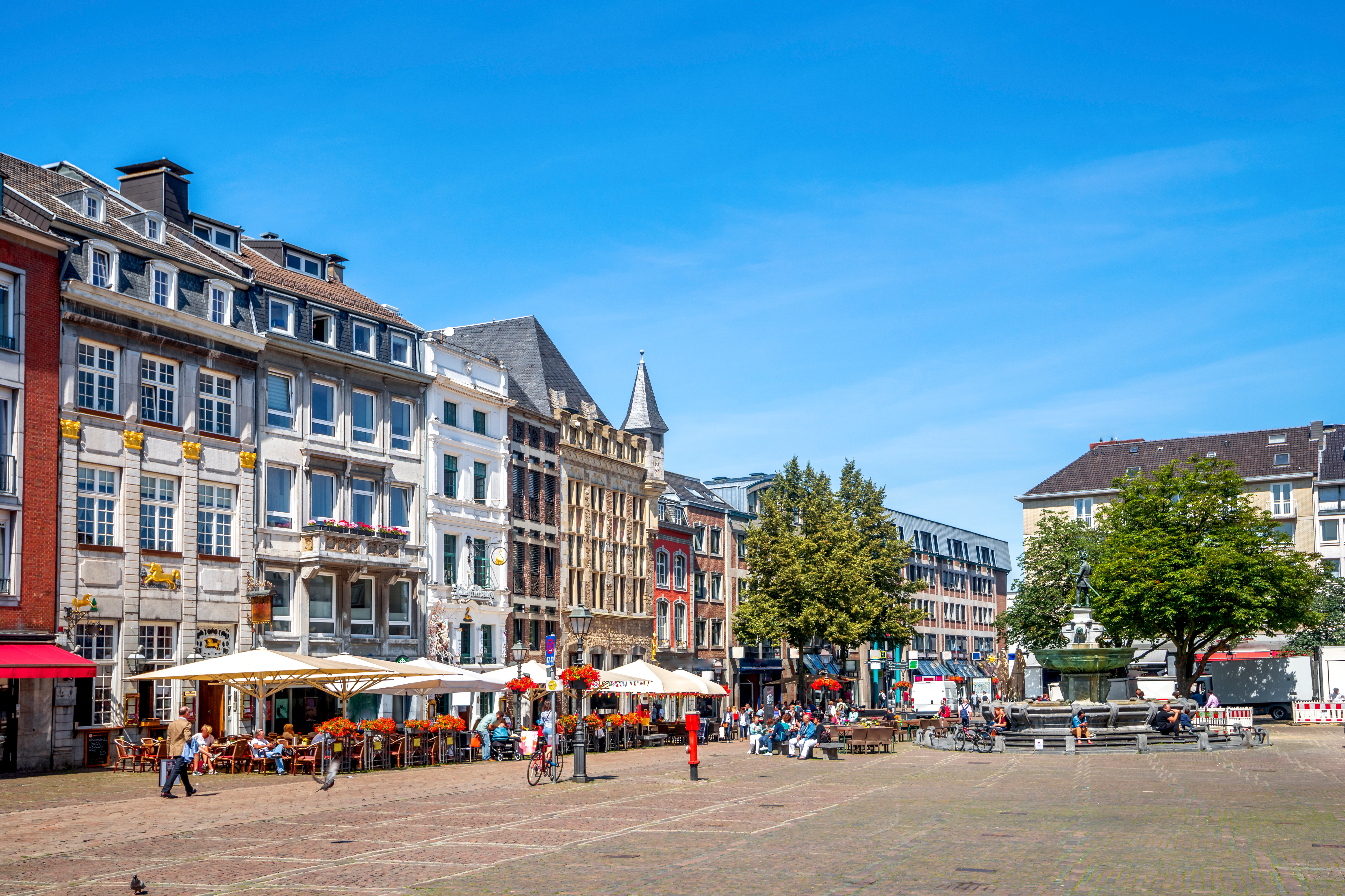 Blick auf Platz mit historischen Wohngebäuden, Straßencafés und Bäumen bei blauem Himmel.
