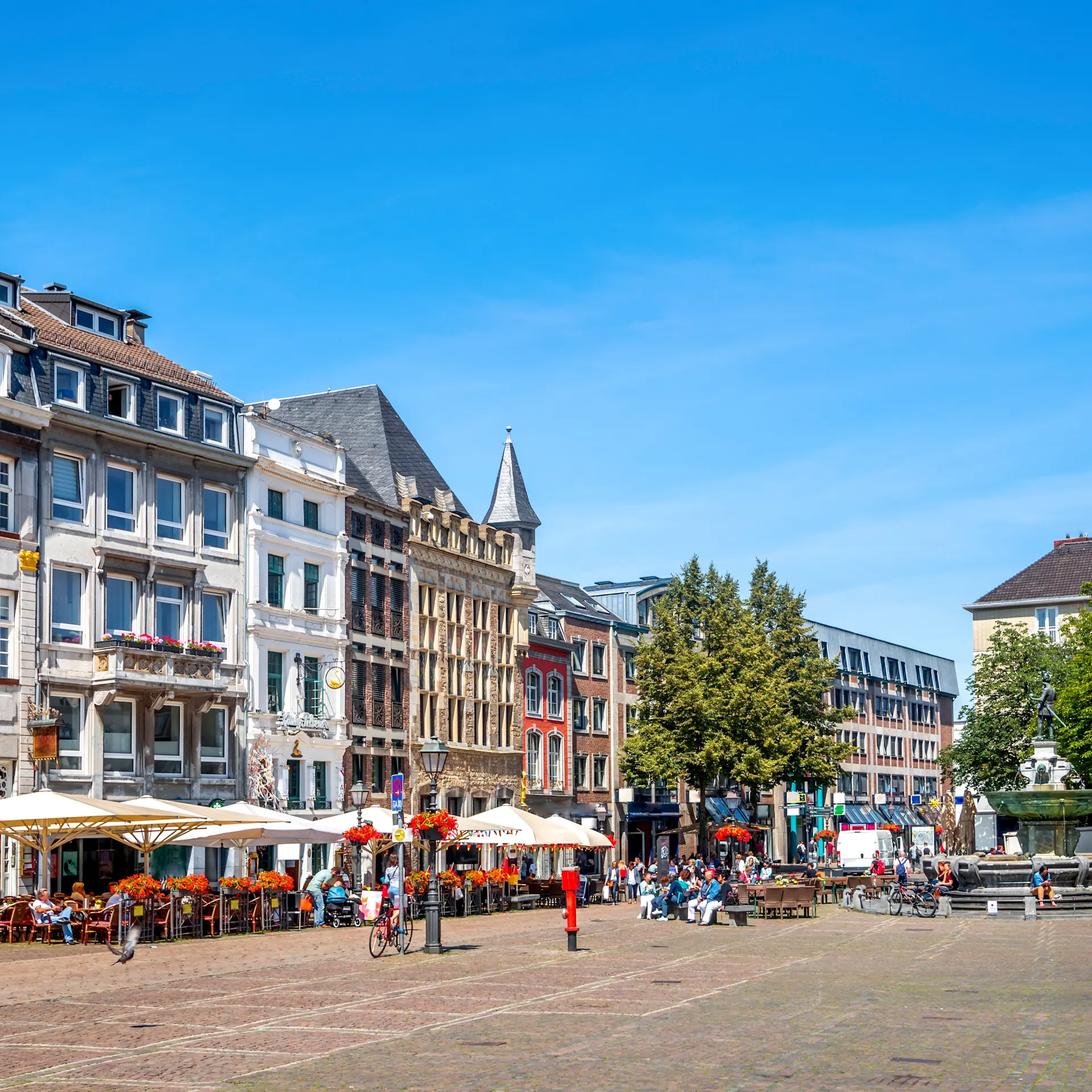 Blick auf Platz mit historischen Wohngebäuden, Straßencafés und Bäumen bei blauem Himmel.