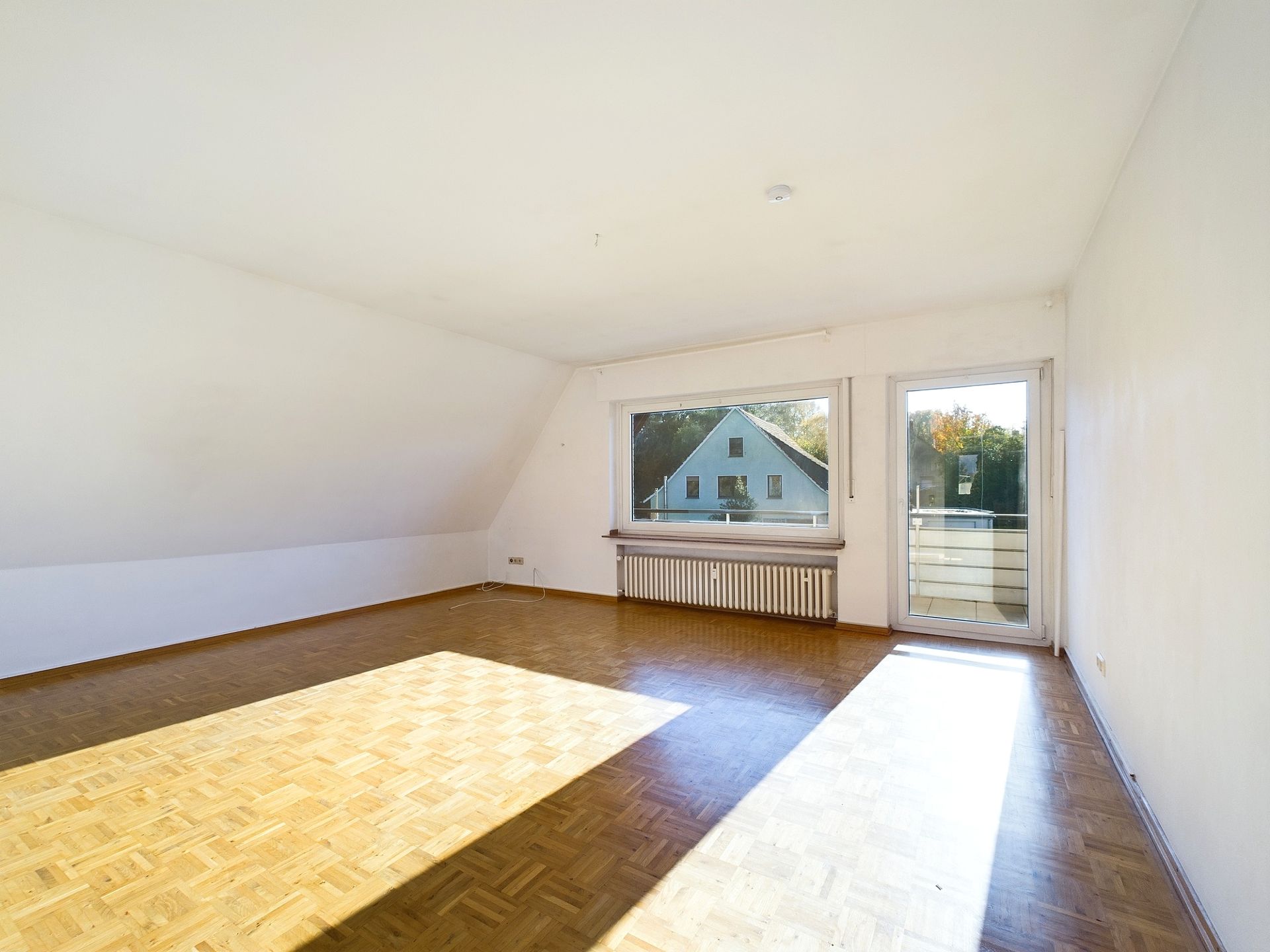 Heller, leerer Raum mit Parkettboden, großem Fenster und Tür zu einem kleinen Balkon.