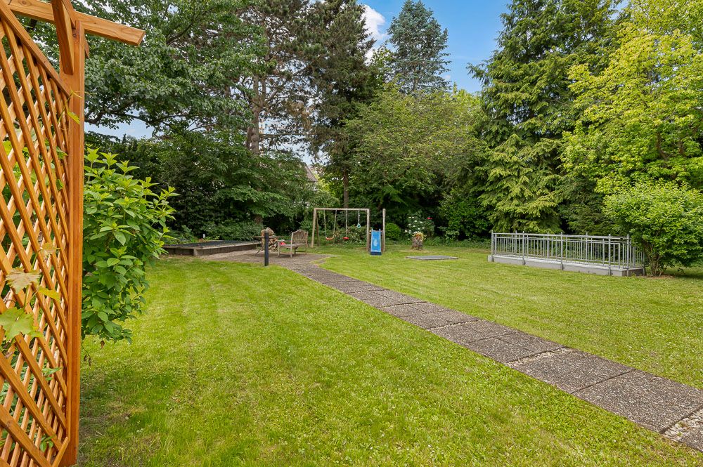 Garten mit Rasenfläche, Gehweg, Spielplatz und Holzgitter, umgeben von Bäumen.