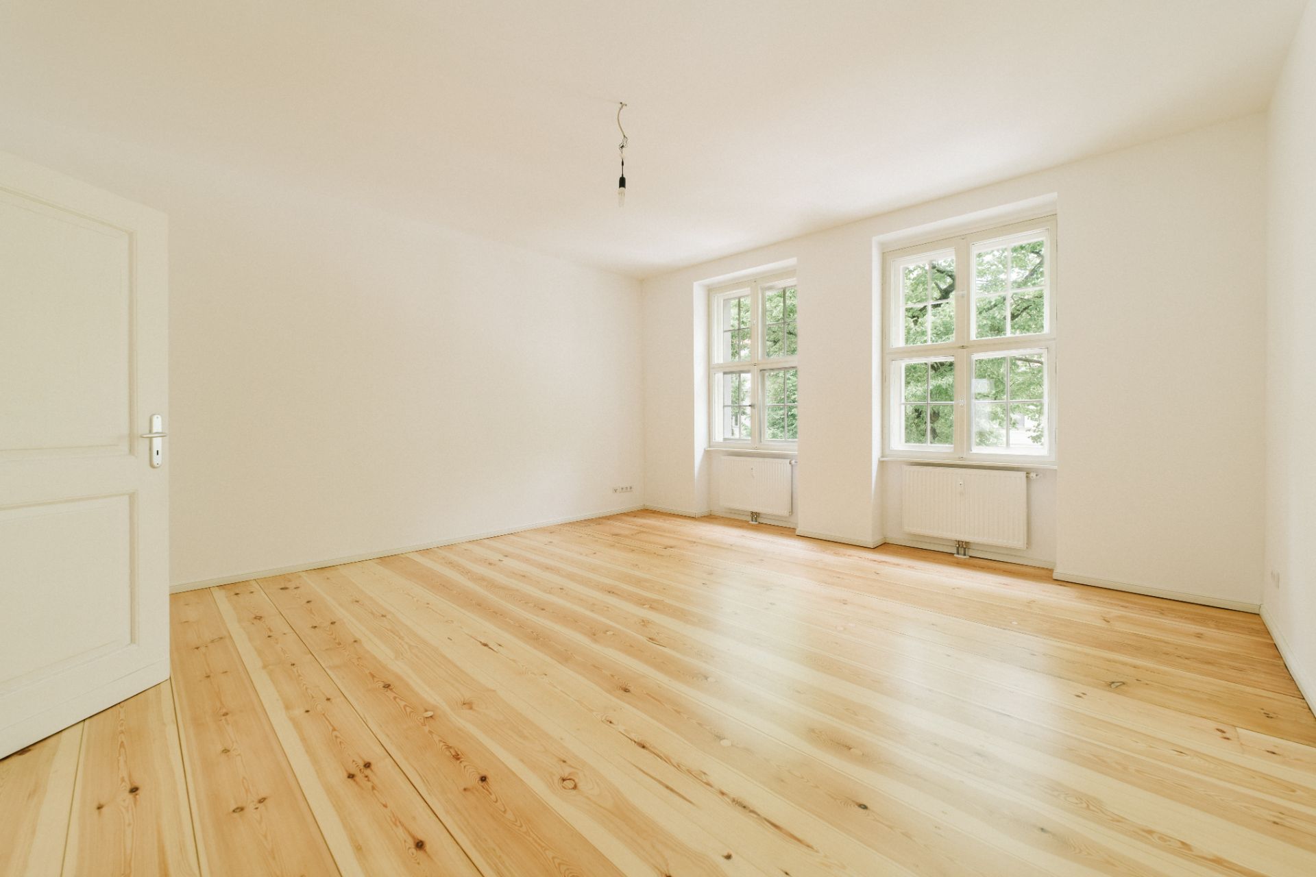 Heller, leerer Raum mit Holzboden, weißen Wänden und zwei großen Fenstern.