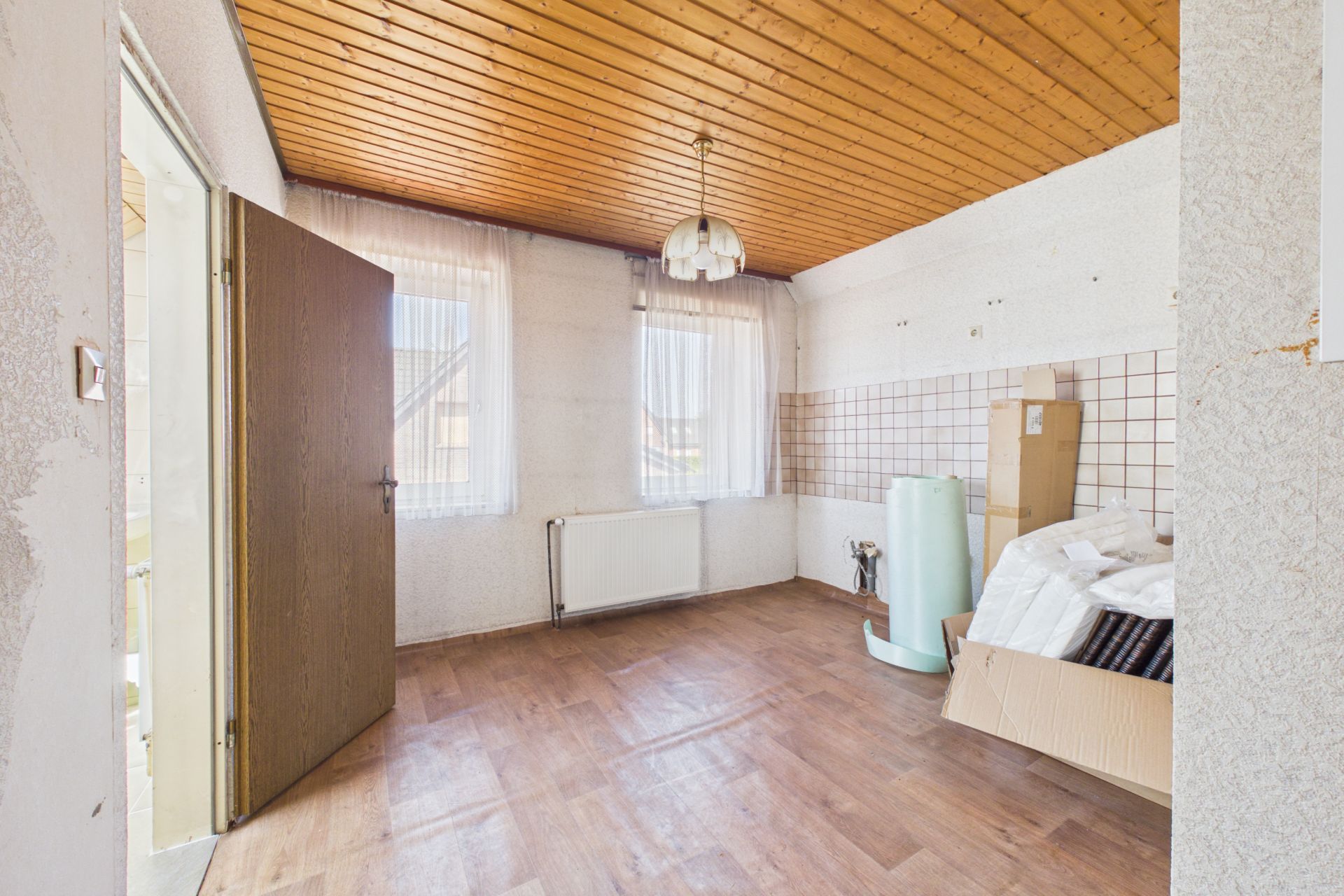 Leerer Raum mit Holzboden, Holzdecke, zwei Fenstern, offener Tür und einigen Kartons an der Wand.