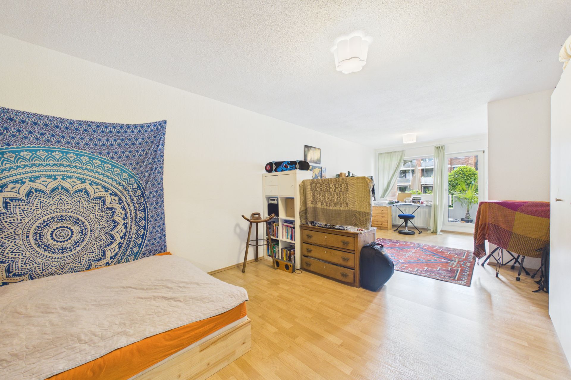 Helles Zimmer mit Bett, Kommode, Teppichen, Regalen und Zugang zu einem Balkon.