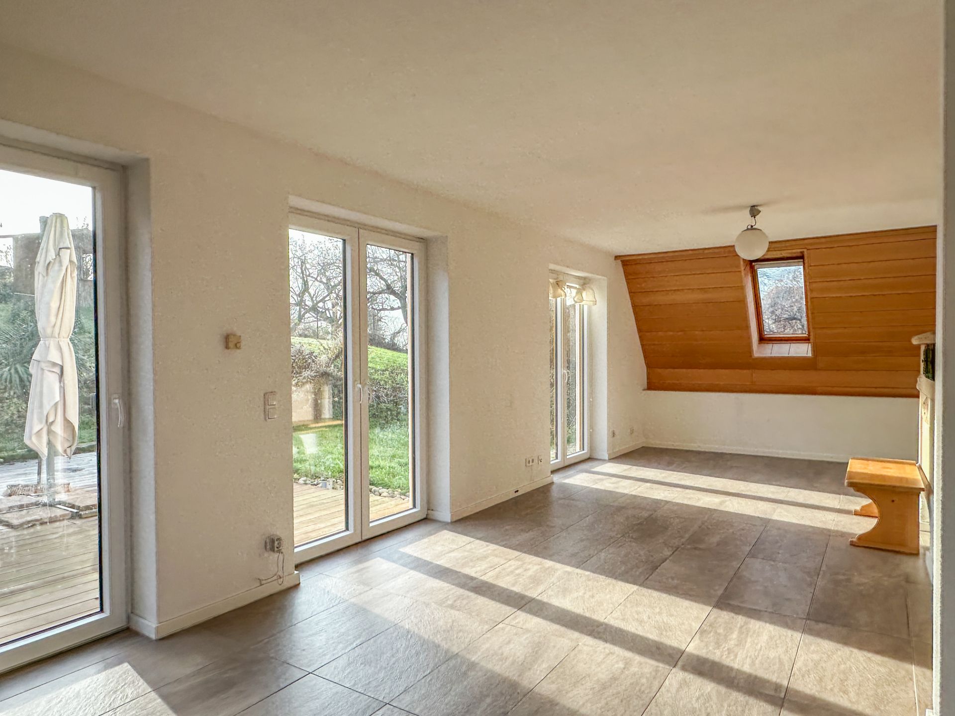 Heller Wohnraum mit großen Fenstern, Fliesenboden und Holzbank, Blick auf Garten.