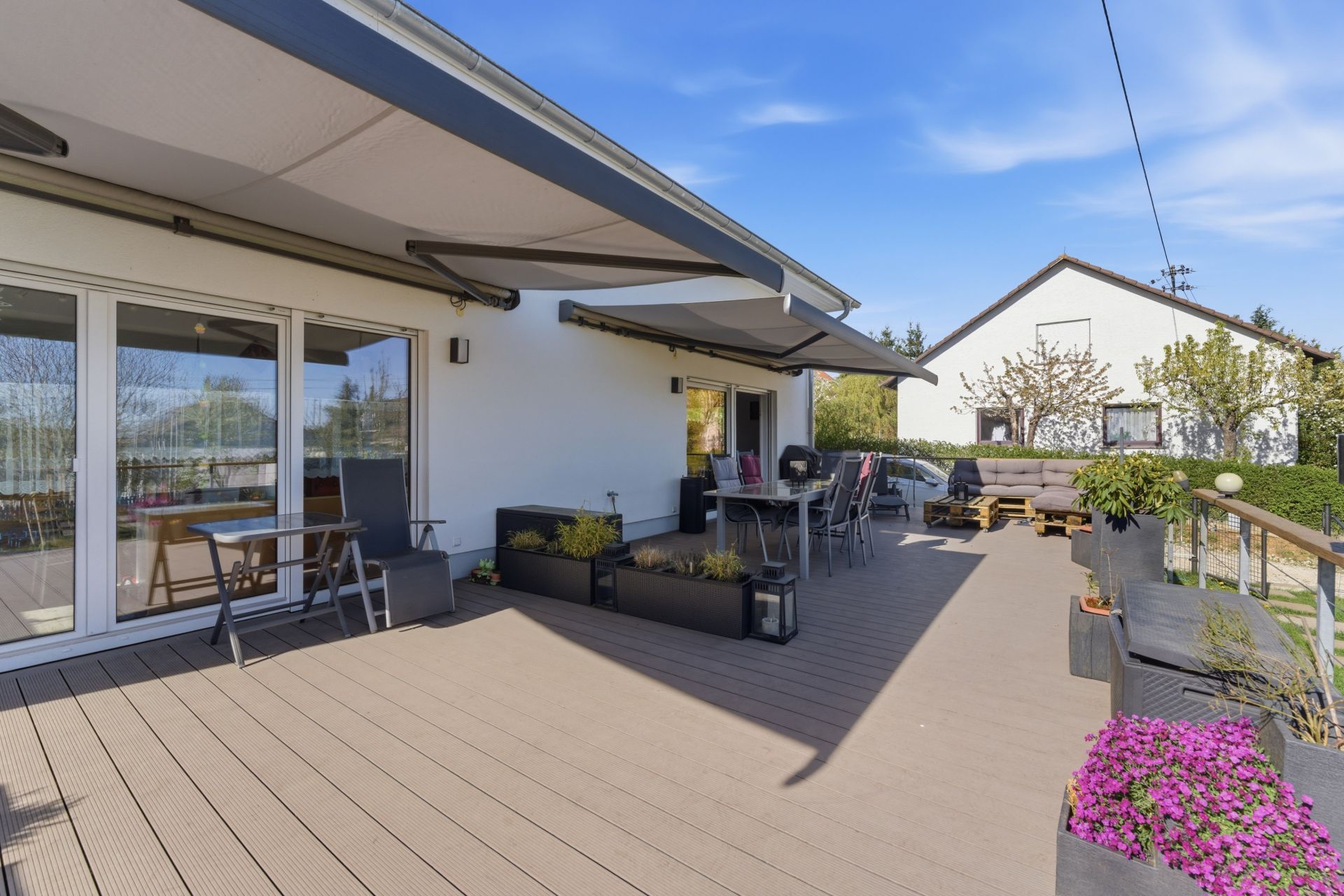 Große Terrasse mit Markisen, Gartenmöbeln, Blumentöpfen und Blick auf einen Garten.