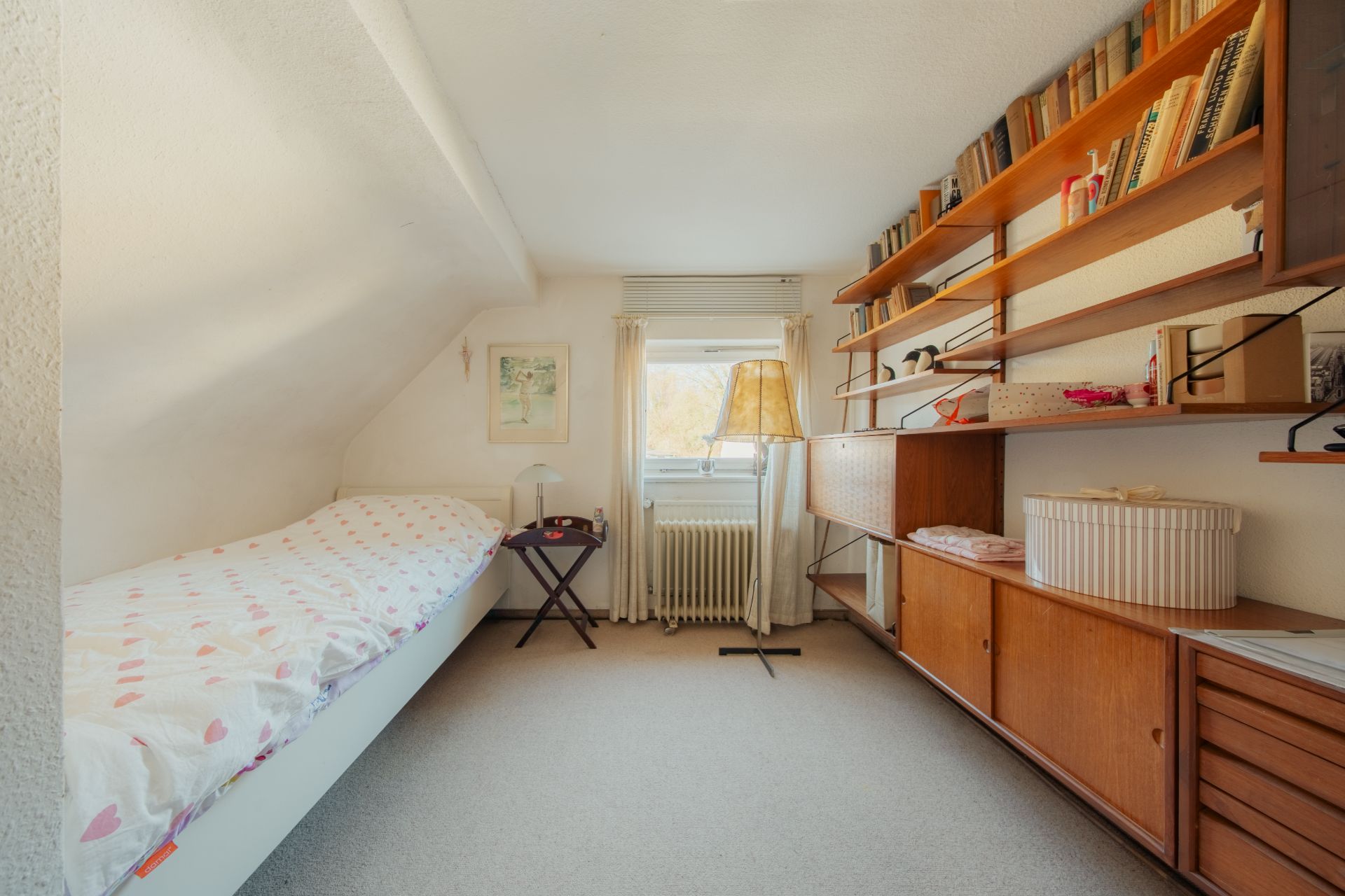 Kleines Zimmer mit Einzelbett, Regalwand, Fenster und Stehlampe.