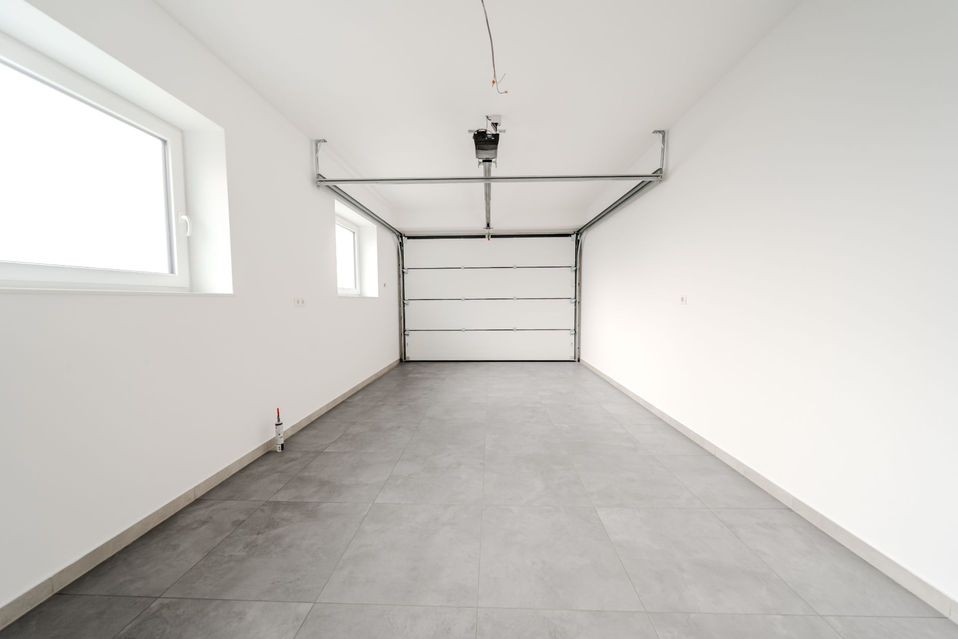 Leere Garage mit grauem Fliesenboden, zwei Fenstern und geschlossenem Garagentor.