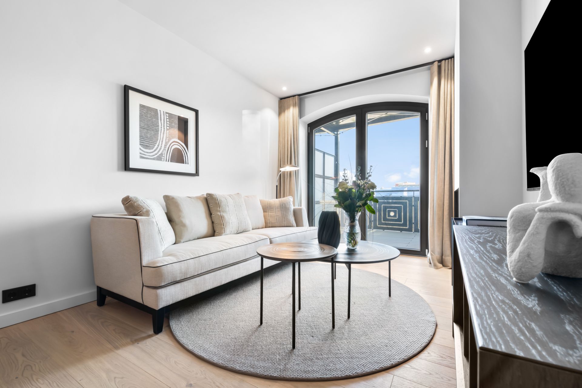 Helles Wohnzimmer mit Sofa, runder Teppich, zwei Tischen und großem Fenster mit Vorhängen.