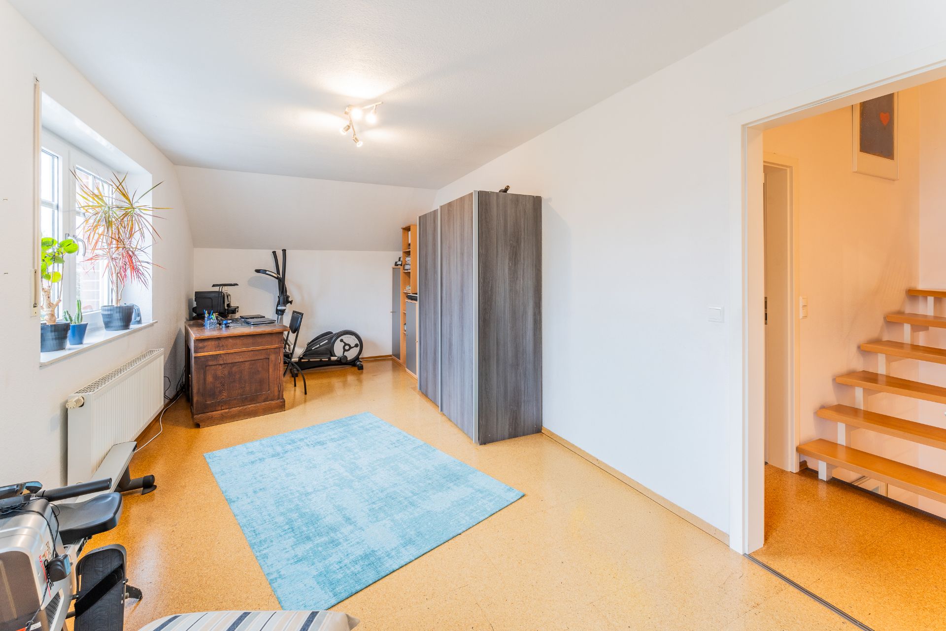 Helles Zimmer mit Schreibtisch, blauem Teppich, Sportgerät und Regal, daneben eine Treppe.