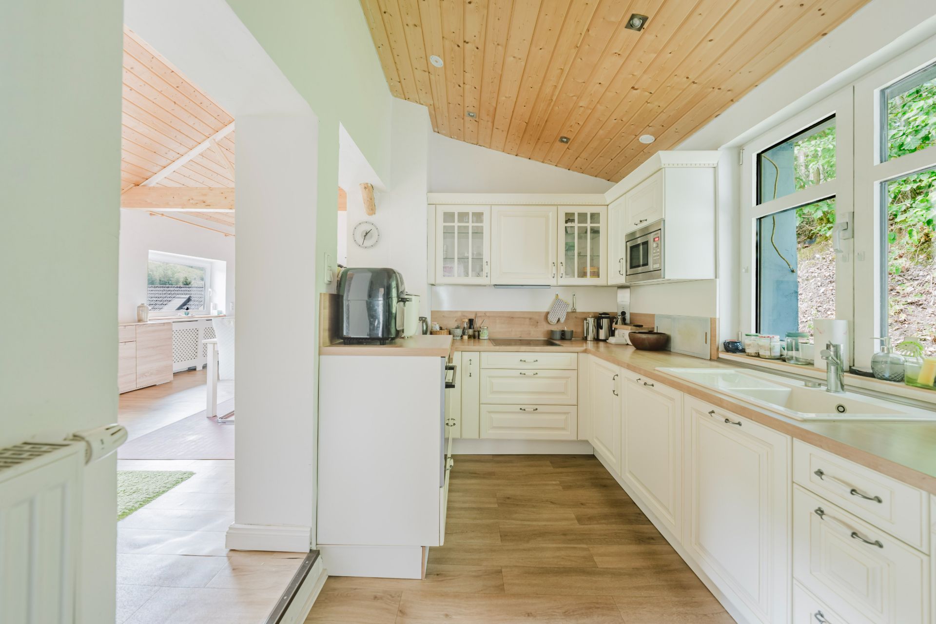 Helle Einbauküche mit weißen Schränken, Holzboden und Fenstern mit Blick ins Grüne.