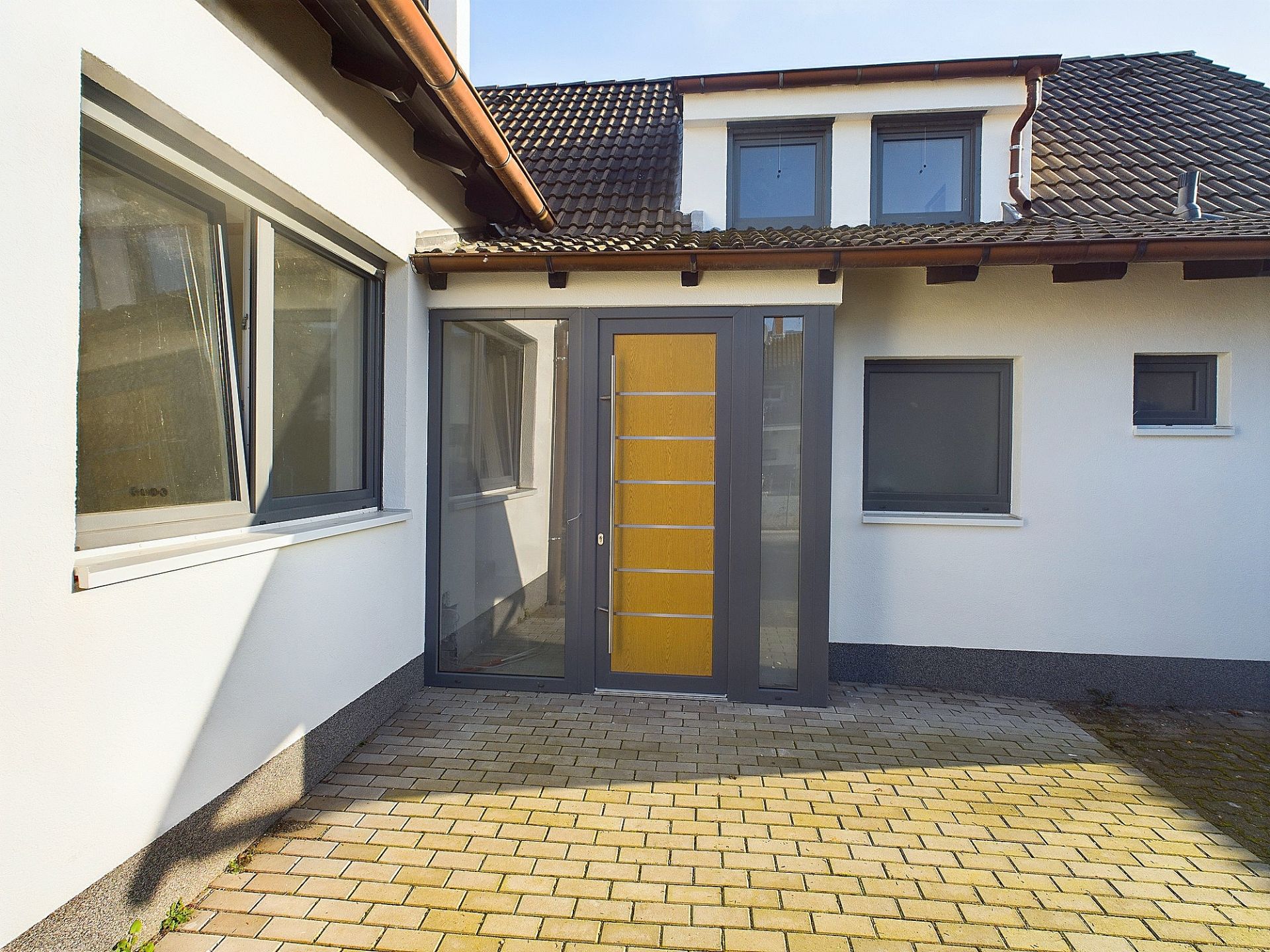 Eingangsbereich eines Hauses mit gelber Tür, großen Fenstern und gepflastertem Hof.