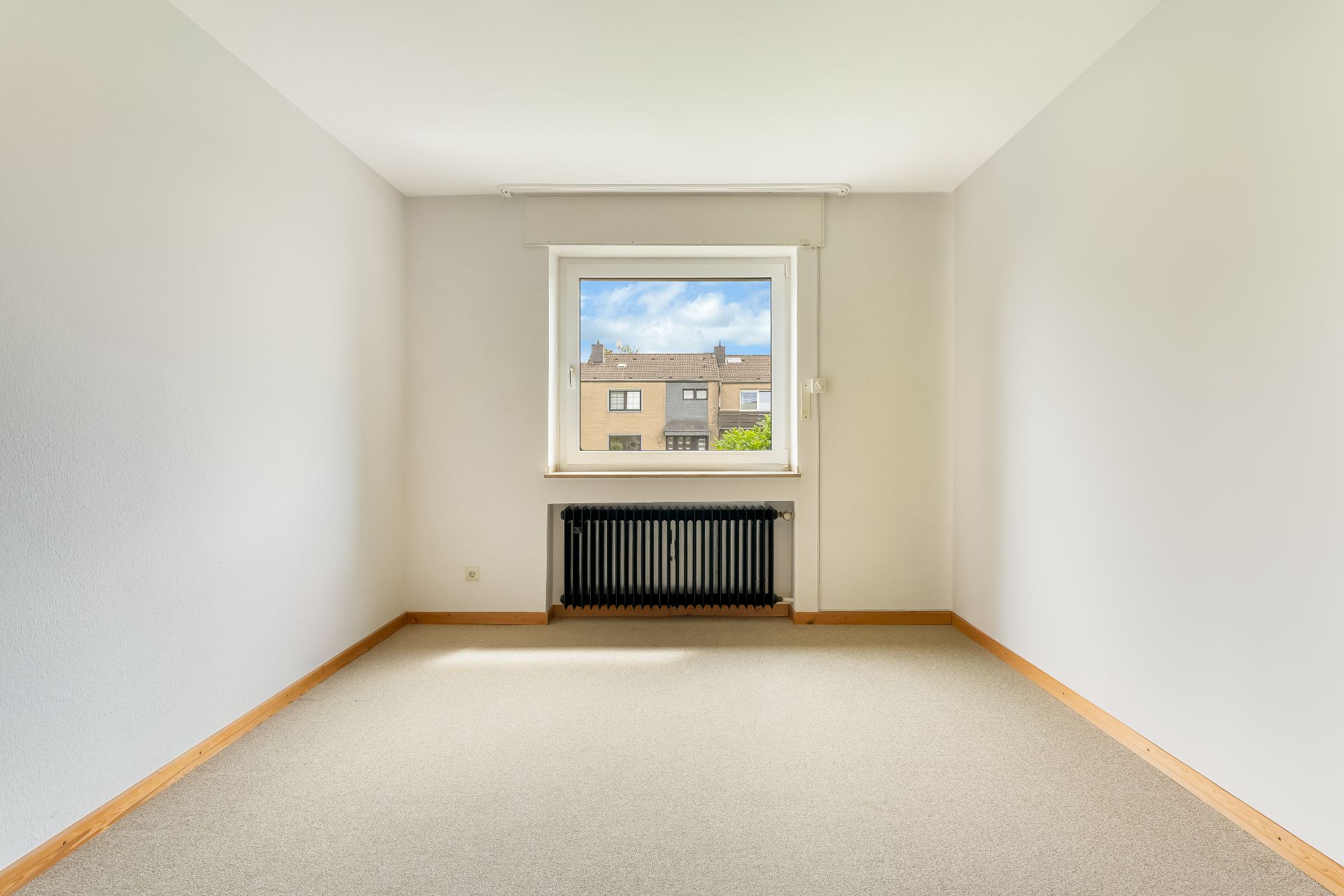 Leerer, heller Raum mit Teppichboden, Fenster und schwarzem Heizkörper unter dem Fenster.