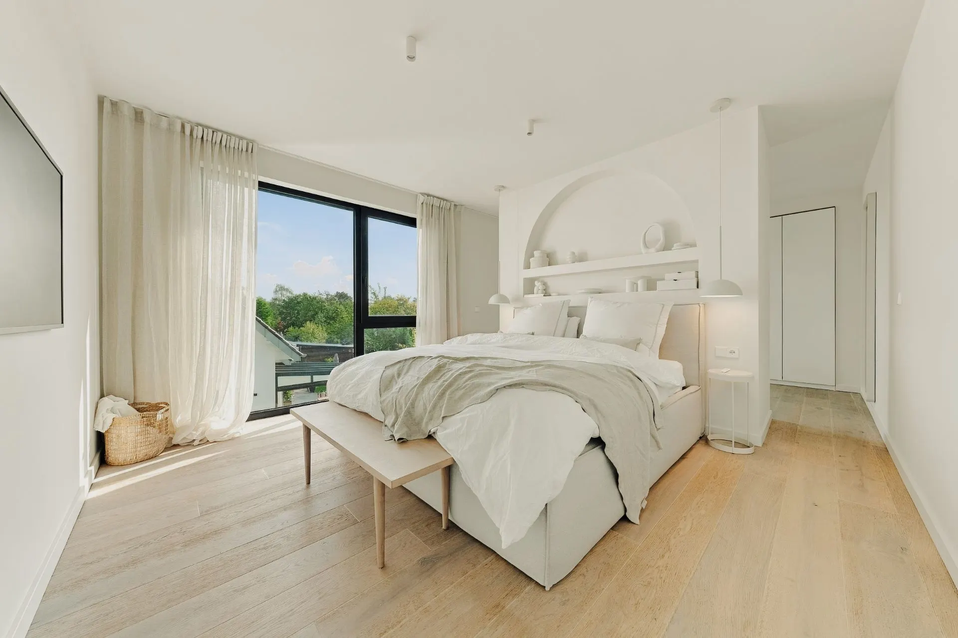Helles Schlafzimmer mit großem Bett, Holzfußboden und breitem Fenster mit Blick ins Grüne.
